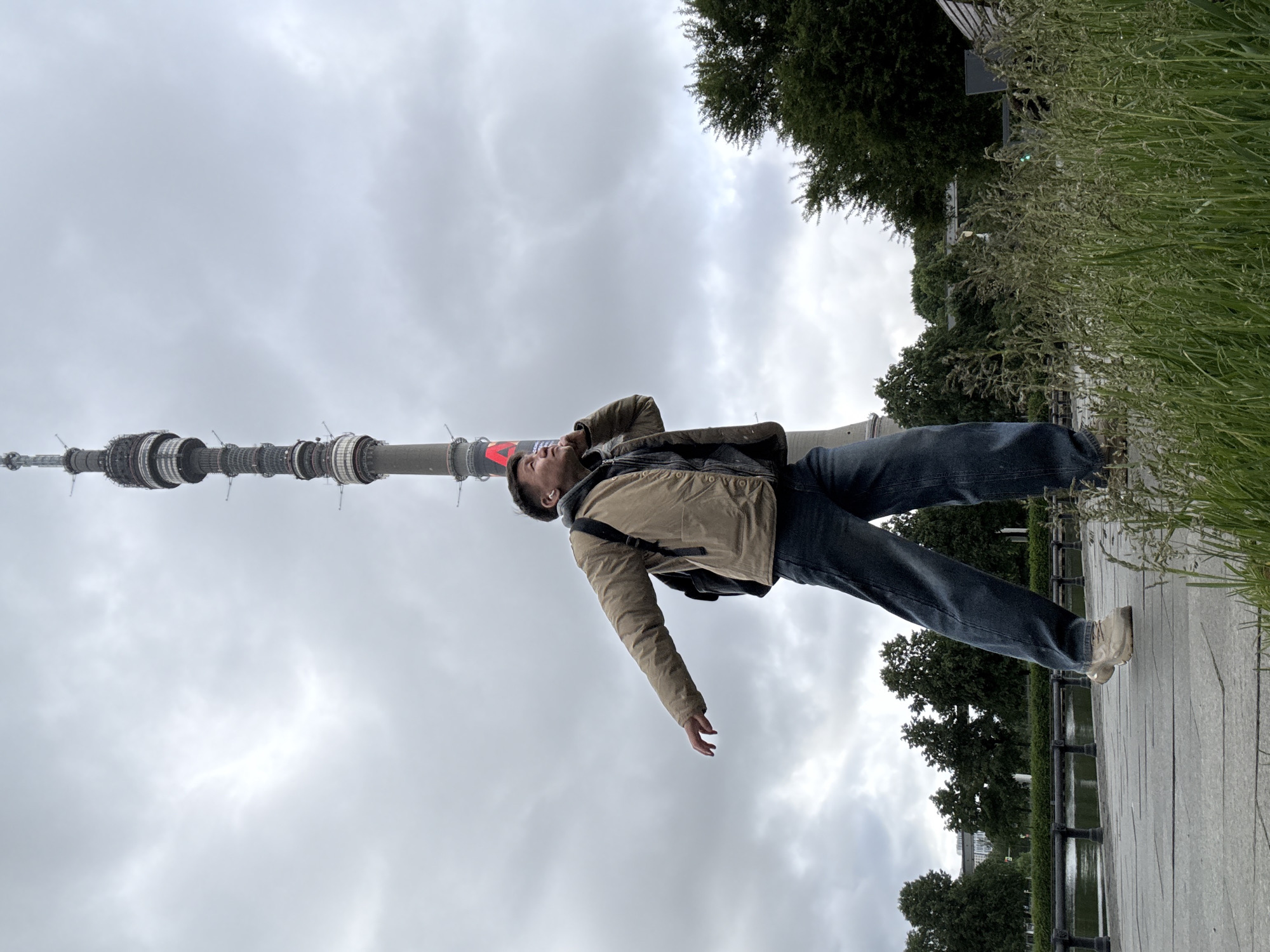 A man wearing a beige jacket, dark blue jeans, and light-colored shoes is lying on a paved surface near a body of water, possibly a pond. Behind him is the Ostankino Tower, a tall television and radio tower in Moscow, Russia, indicating the image was taken near that location, which is in the city of Khimki, Russia. Trees and greenery surround the paved area. The sky is overcast.