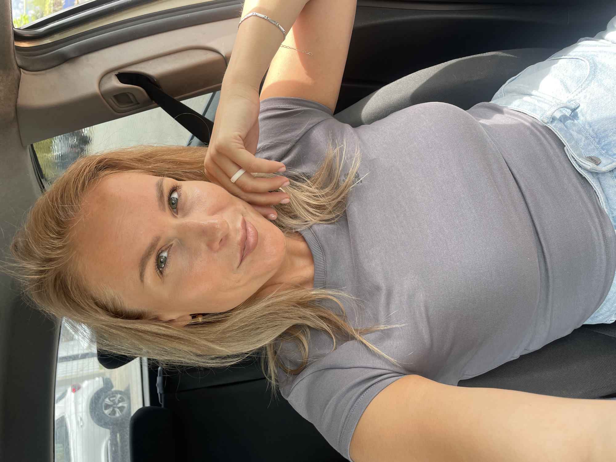 A blond-haired woman is sitting in a car, possibly a vehicle, with the interior visible. She is wearing a gray t-shirt and a light blue denim jacket. She has a ring on her finger and a bracelet on her wrist. Through the car window, another vehicle can be seen, along with an outdoor environment. There is no explicit indication of the location being Sochi, Russia in this image.