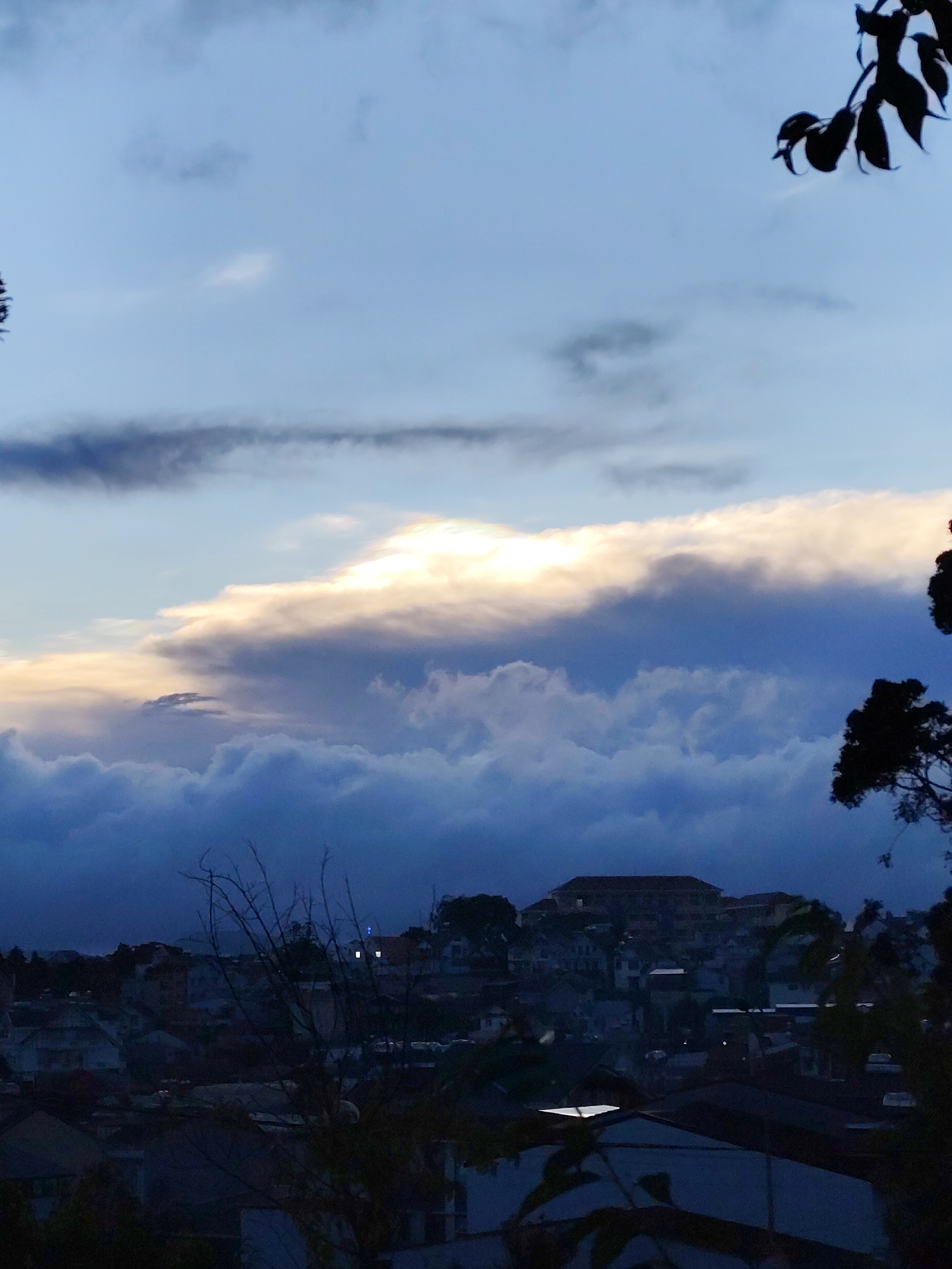 The image shows a view of Lang Biang - Da Lat, Vietnam at dusk. In the foreground, silhouetted trees and rooftops are visible, indicating an elevated perspective. The middle ground features buildings and houses nestled among trees, creating a sense of urban density. The background is dominated by a dramatic sky with a mix of dark and light clouds, illuminated by a partially obscured sun. The overall atmosphere is one of tranquility, with a focus on the natural beauty of the sky contrasted with the urban landscape.