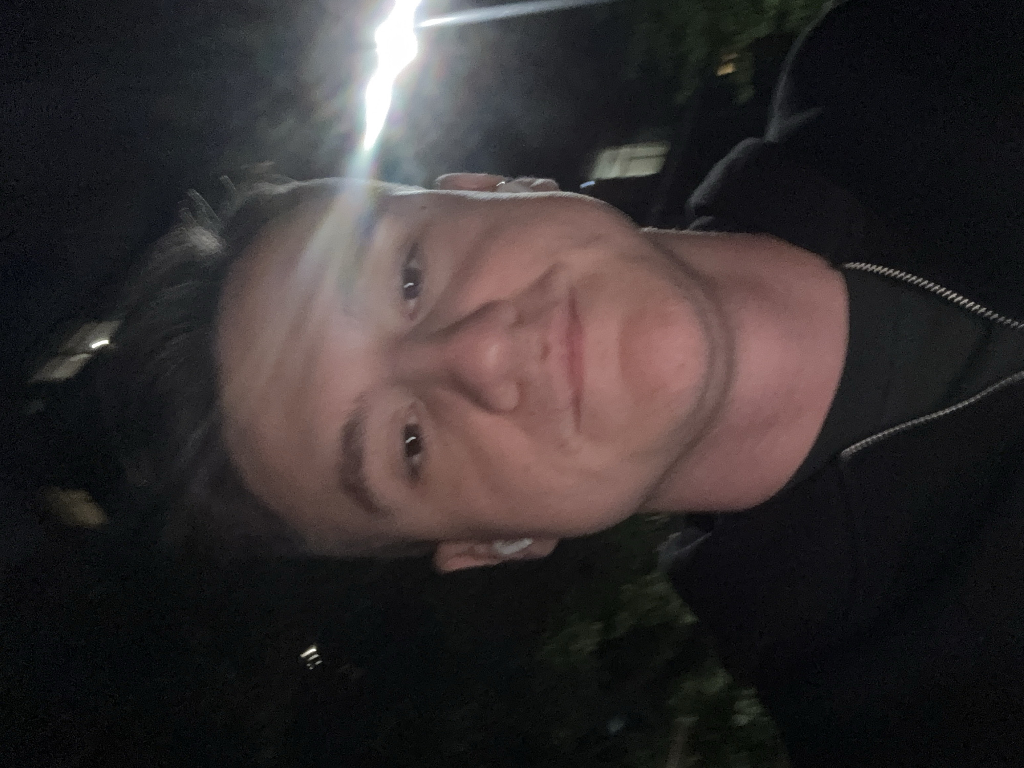 The image is a close-up self-portrait of a person with fair skin. The person has short, dark hair and is wearing what appears to be a dark-colored hooded sweatshirt or jacket. A silver chain is visible around their neck. An earpiece is present in their ear. The background is dark and appears to be outdoors at night, with some foliage and a bright light source visible in the distance. The location in the city is unknown, unknown.