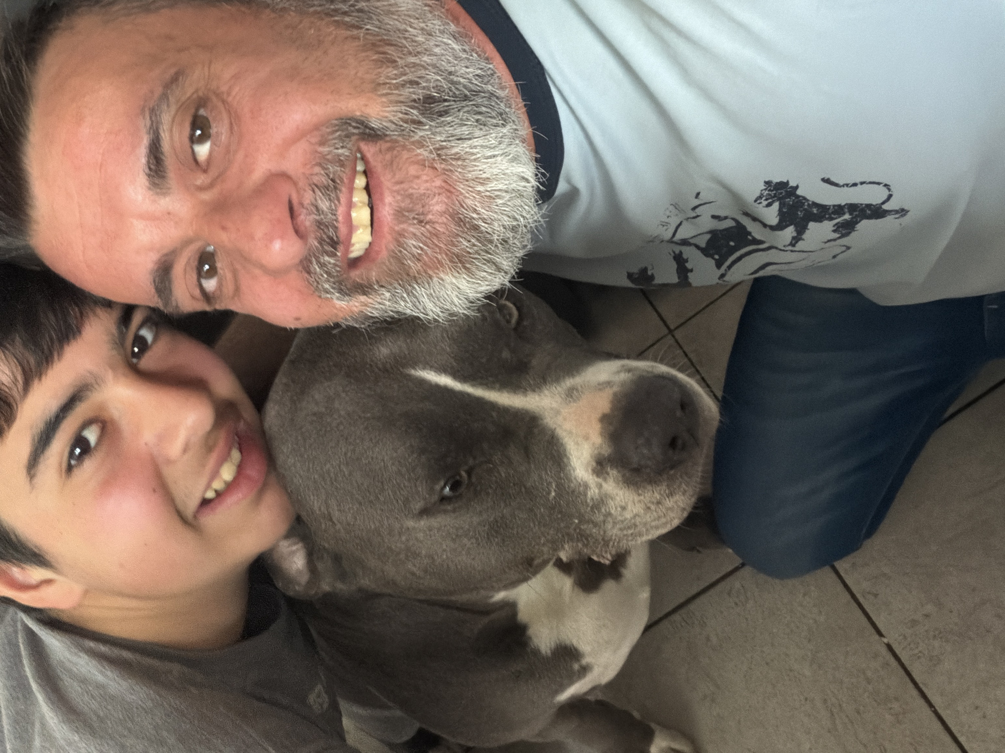 A selfie-style image shows an adult male, a young male, and a pit bull-type dog. The adult male is positioned at the top of the frame, with graying hair and a beard. He is wearing a light blue t-shirt with a dark blue collar, which features a black graphic of a lion-like figure. He is smiling, revealing his teeth. The young male is positioned on the left side of the frame, wearing a gray shirt. He is also smiling. The pit bull-type dog is positioned in the center, with its head resting between the two males. The dog is primarily gray, with white markings on its face and chest. The floor is tiled in a neutral tone. The location cannot be determined based on the visual data provided.