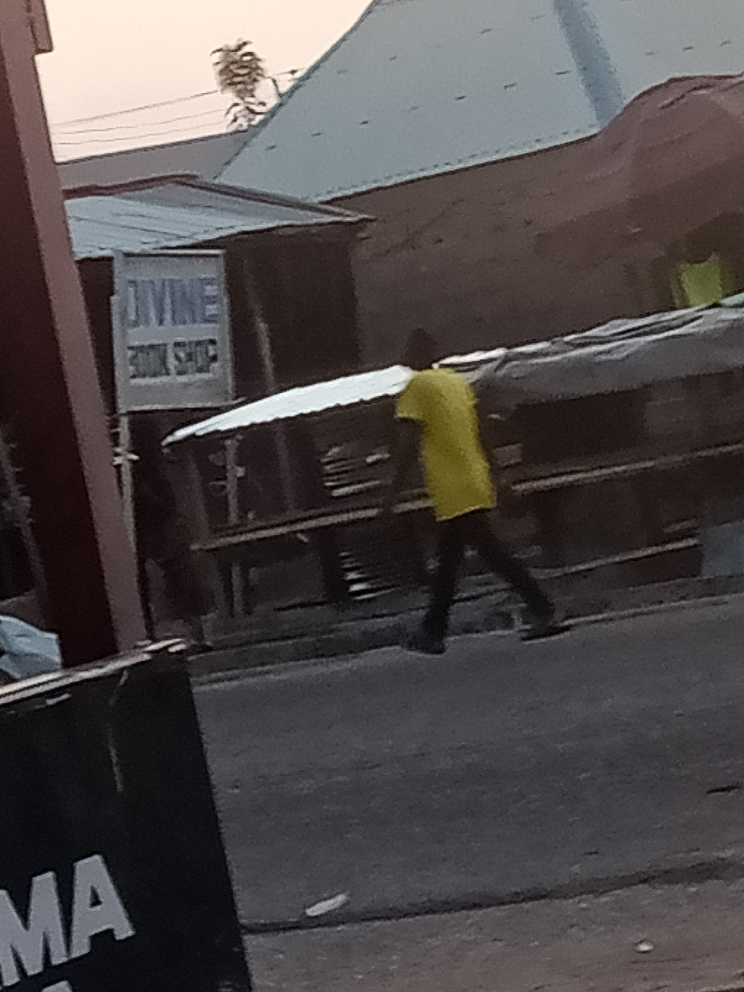 The image depicts a street scene in Jalingo, Nigeria. A person wearing a yellow t-shirt and black pants is walking on the road. Behind them, there is a structure with a sign that reads "DIVINE BOOK SHOP". Buildings and structures associated with a market or commercial area are visible in the background. The lighting suggests it might be late afternoon or early evening. A partial view of a black sign with white lettering, possibly advertising a business, is visible in the foreground on the left side of the image.