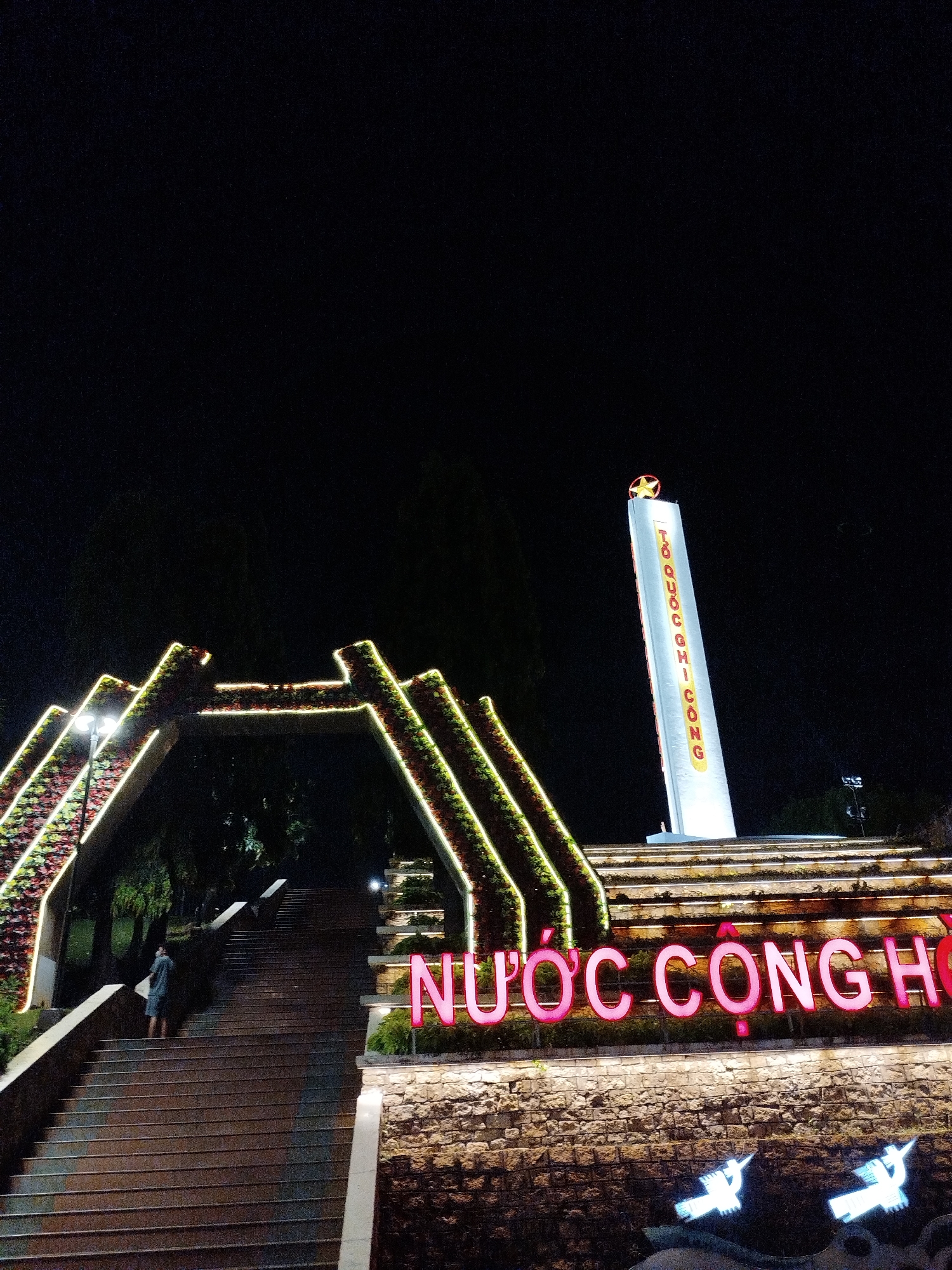 At night, a man is walking up a long staircase at the historical tourist attraction, the Independence Palace (Reunification Palace), located in Ho Chi Minh City, Vietnam. The entrance to the stairs has an angular floral archway with lights. In the background is a large, white monument topped with a red star. In the foreground, red neon lights spell out the Vietnamese words "Nước Cộng Hòa". Below that, white neon lights depict two birds in flight. The sky is dark.