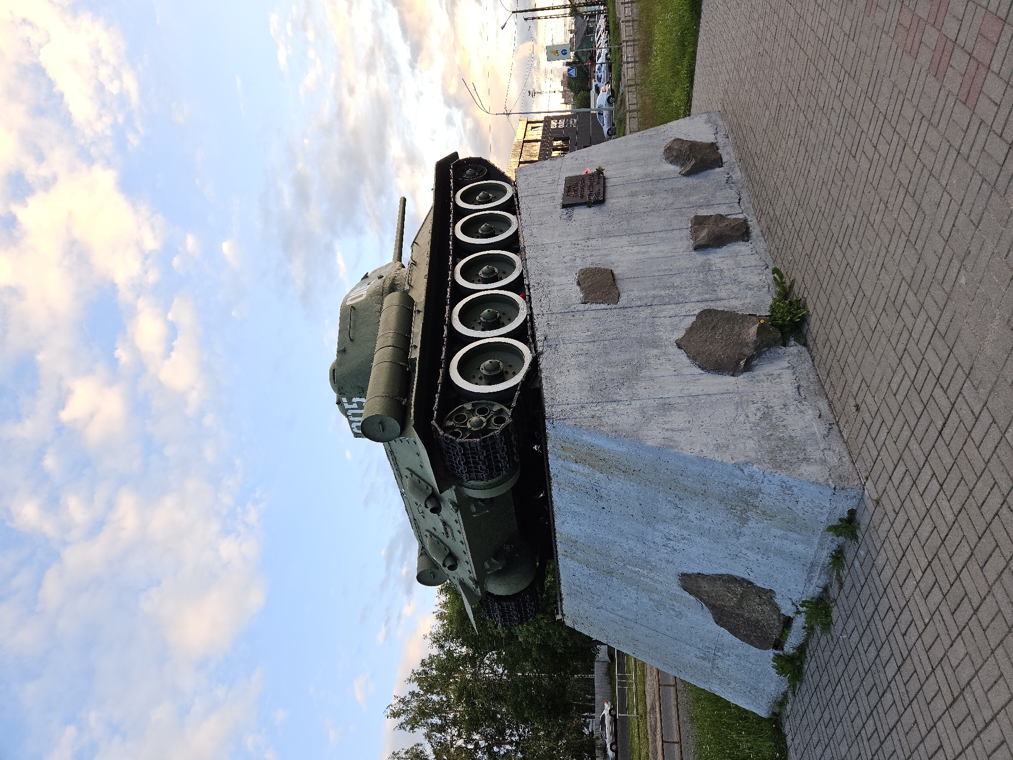 This image features a preserved military tank displayed as a monument. The tank, painted in a dull green, is positioned on a concrete pedestal. Its tracks and road wheels are clearly visible. The pedestal itself is rough and textured, with several large stones embedded in its surface. To the right of the pedestal, a brick-paved walkway is visible, suggesting an outdoor public space. Beyond the walkway, there is a patch of green grass and trees. In the background, there are signs of urban life, including streetlights, buildings, and parked vehicles, indicating the monument is located in a town or city. The sky is partly cloudy with soft, warm light, suggesting it might be late afternoon or early evening. There is a plaque attached to the concrete pedestal, but the text is not legible. The overall impression is that of a historical memorial site.