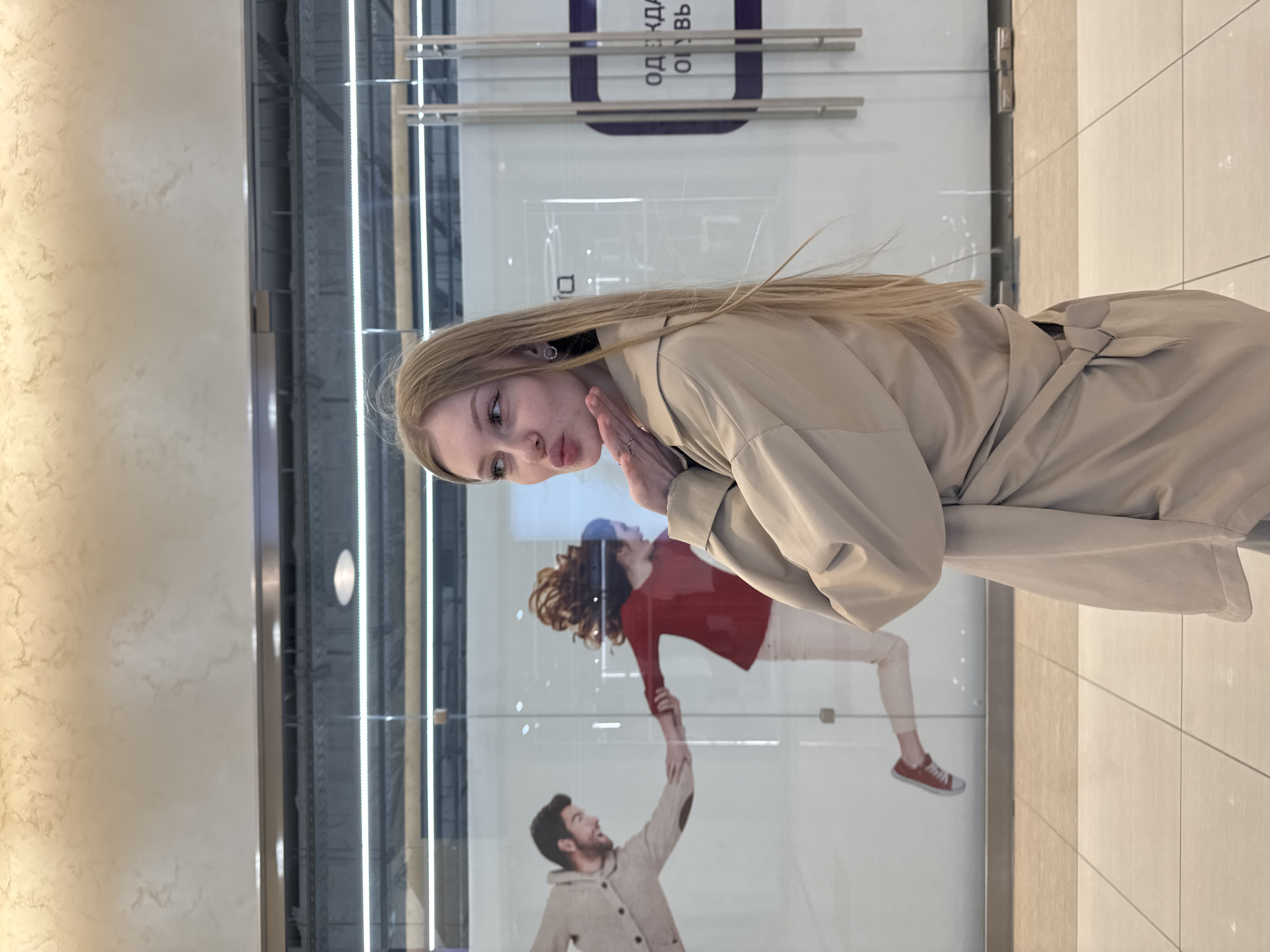 A woman stands in front of a storefront window. The woman is Caucasian with long blonde hair, wearing a tan trench coat. She is posed as if blowing a kiss.
Behind her is a storefront window with a promotional image. In the image, a man reaches up toward a woman who is suspended in the air; the man wears a tan cardigan and the woman wears a red long-sleeved shirt with white pants and red tennis shoes.
The storefront sign above the promotional image says "ОДЕЖДА ОБУВЬ" which translates to "CLOTHING SHOES". The flooring appears to be a beige tile.
Based on online searches, the Cyrillic text on the store sign, the interior design of the storefront, and the woman's attire, the location is likely a shopping center in Perm, Russia.