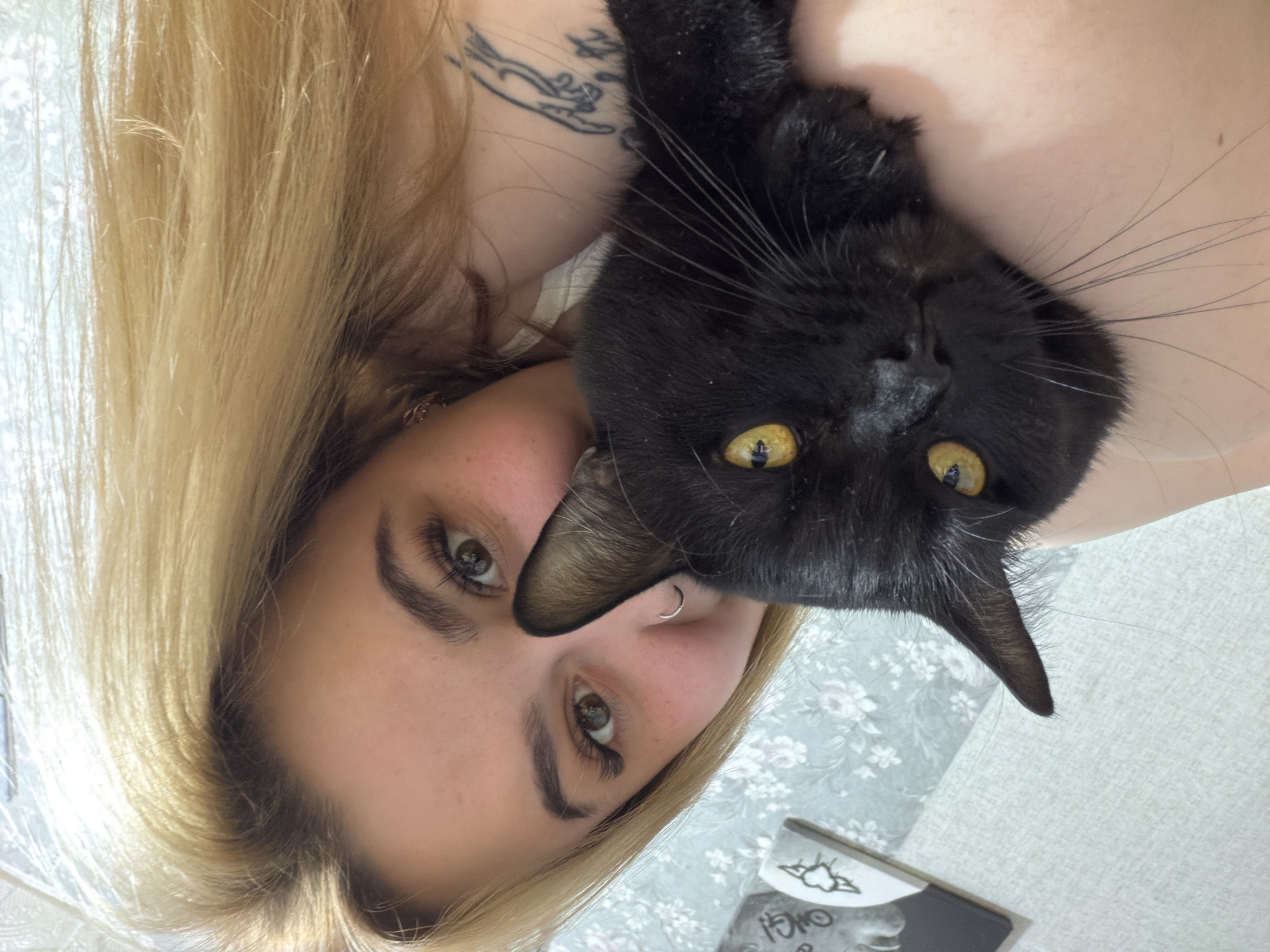 A fair-skinned woman with blonde hair is holding a black cat. The cat's head is positioned near the woman's face. The woman has a nose ring and a tattoo is visible on her upper chest/shoulder area. The background appears to be a floral pattern. The presence of a document with the city name "Chaikovsky" in Cyrillic is suspected from the crop images. Therefore, the location is presumed to be Chaikovsky, Russia.