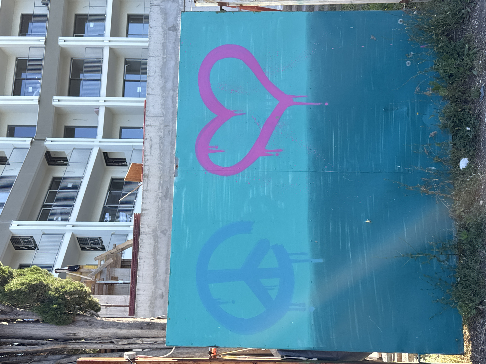 The image presents a scene in Rafailovići, Montenegro, dominated by a large, teal-colored painted board situated next to a multi-story building under construction. The board features a pink heart-like symbol in its upper half and a blue peace symbol below. The teal paint has drip marks indicating it was spray painted. The building, constructed of concrete and glass, has a modern design with multiple balconies. Construction materials and wooden supports are visible at the base of the building. To the right of the board is a strip of vegetation. The image is taken from a medium distance with a slight upward angle, capturing the juxtaposition of the colorful graffiti with the ongoing construction.