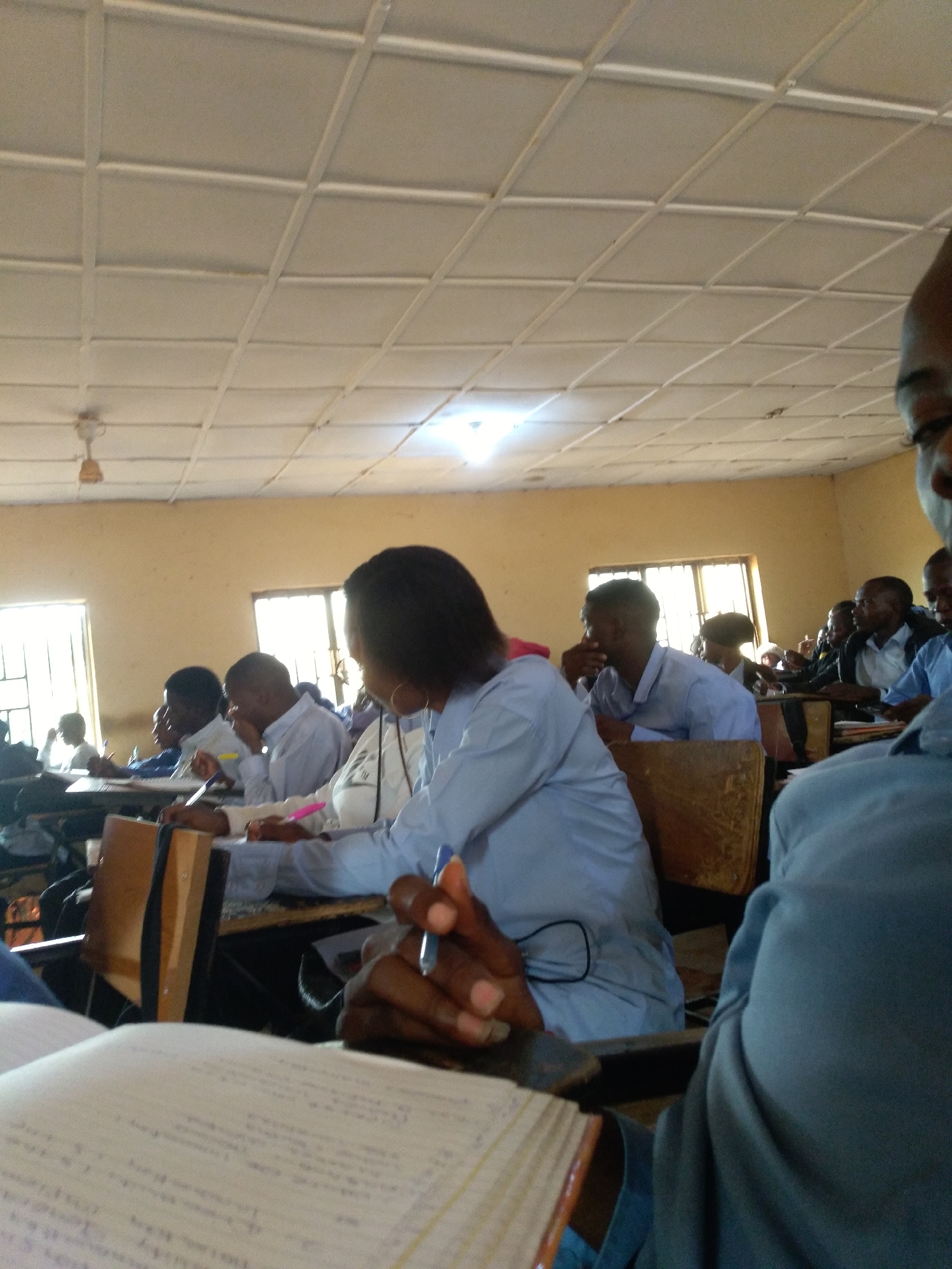 This image depicts a classroom scene in Jos, Nigeria, likely during the daytime given the natural light filtering through the windows and the bright overhead lighting. The room appears to be a typical educational setting with rows of desks and chairs, occupied by students. The walls are painted a light, warm color, and the ceiling is a grid of white tiles with visible fluorescent lighting. A ceiling fan is also visible, suggesting a warm climate.
The primary subjects are a group of young people, presumably students, engaged in what appears to be a lecture or study session. Several individuals are seen from the side or back, focusing on their notes or the front of the room. Some are holding pens and writing, indicating active participation. In the foreground, a student's hands are visible holding a pen and a notebook, with a portion of a book or notebook in the very front. Another student, wearing a light blue shirt, is seated in the mid-ground, also looking down. Further back, other students are visible, some in light-colored shirts. The overall atmosphere seems attentive and studious. There are no strong emotional cues visible, but the focus of the students suggests a serious academic environment.
Notable details include the clothing worn by the students, which appears to be casual but appropriate for a classroom. The presence of desks, notebooks, and pens are objects indicative of a learning environment. The windows show a glimpse of the outside, though it is not clearly discernible. The scene is well-lit, making it easy to see the subjects. There is no visible text in the image that can be clearly deciphered. The general impression is one of a normal school day in progress.