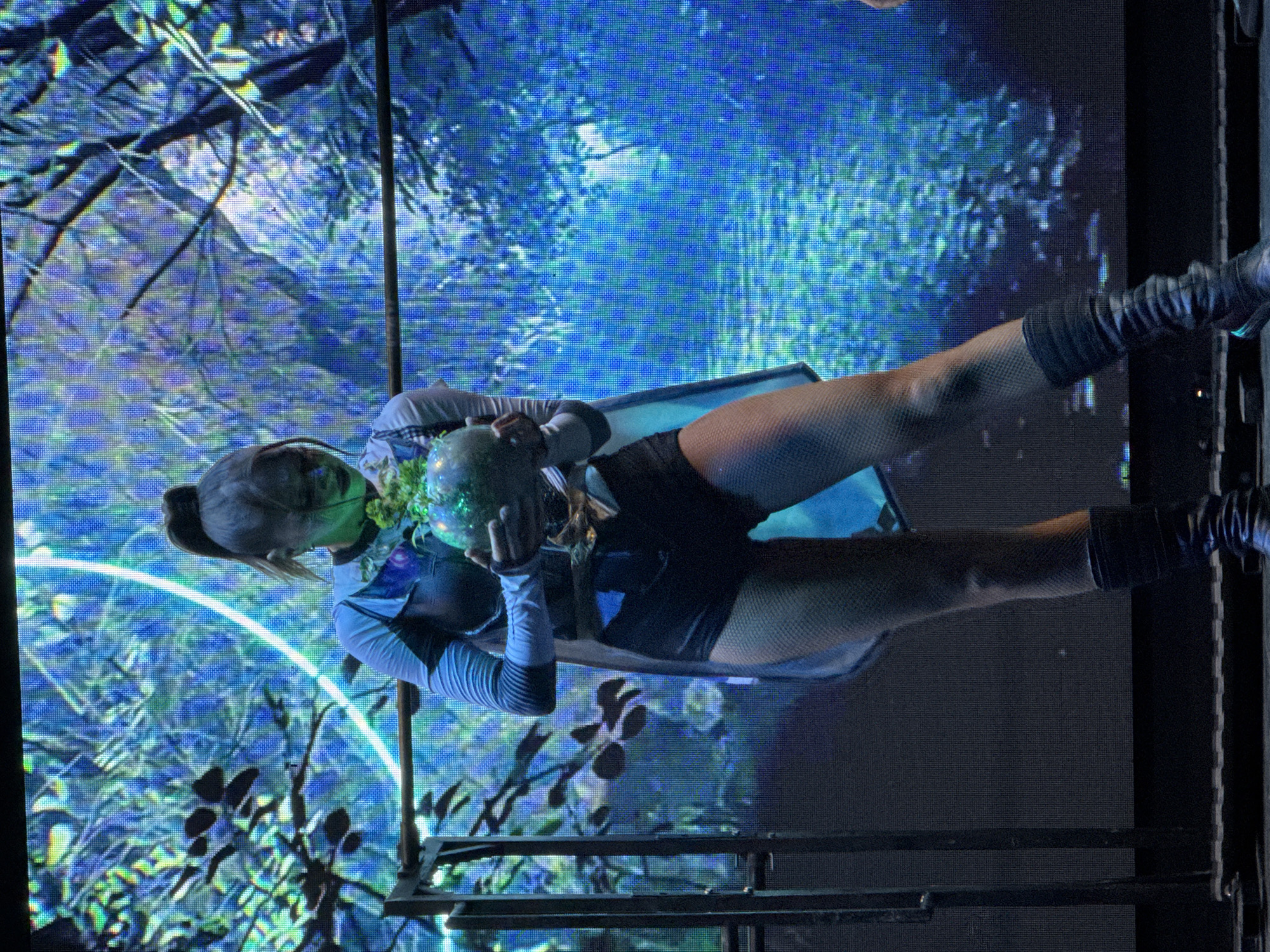 A woman with a ponytail, wearing a blue and black outfit with fishnet stockings and dark boots, stands on a stage, holding a spherical object with green foliage. She is illuminated by spotlights and stands before a large LED screen displaying a vibrant, abstract image of blue and green foliage with a white circular element. The stage floor appears to be black. The image was taken in the city of Manavgat, Turkey.