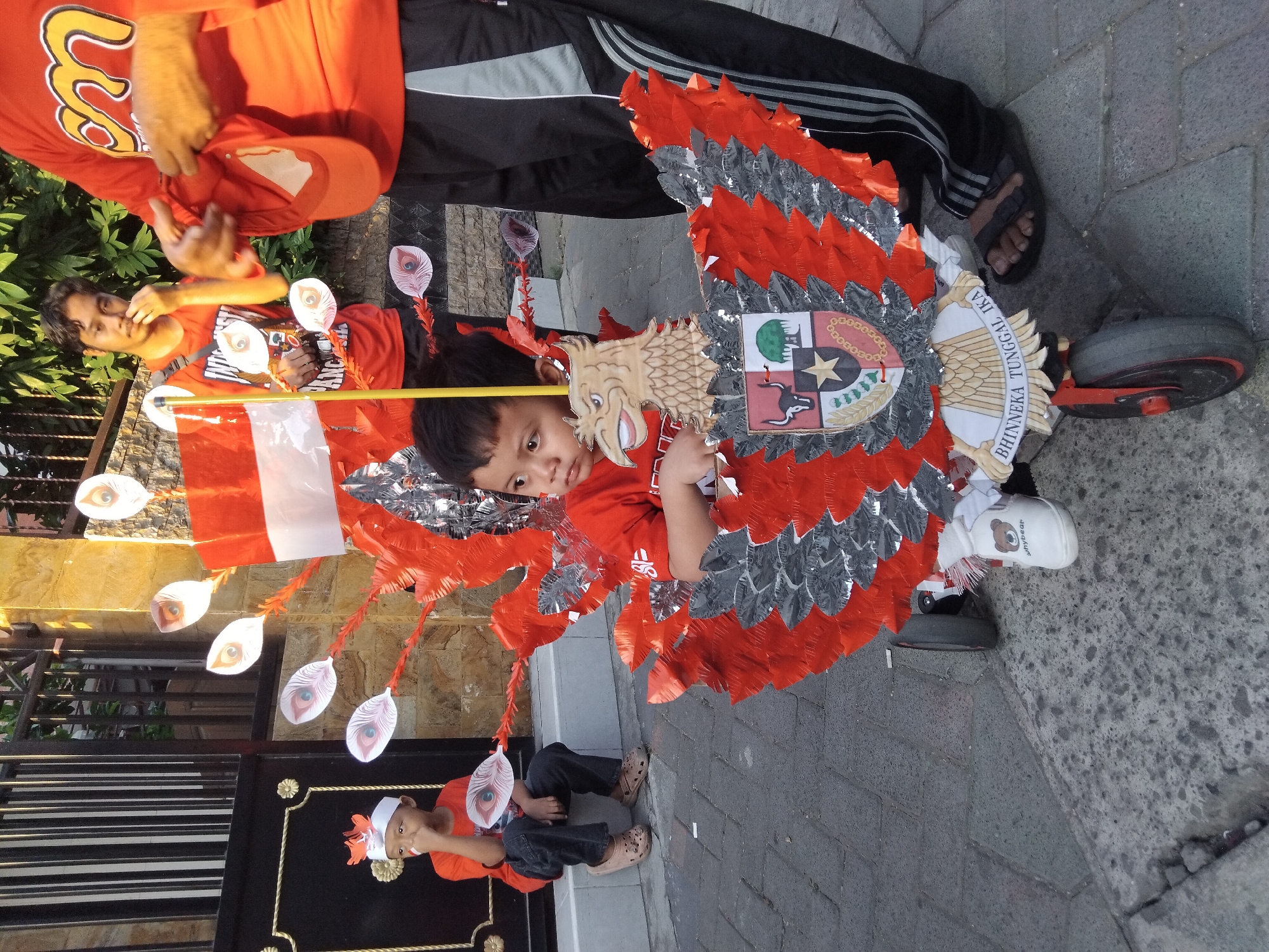 In the city of Kepuh Kiriman, Indonesia, a young boy sits in a wheeled contraption resembling a Garuda bird, which is adorned with red and silver feathers and an Indonesian coat of arms. The boy is wearing a red shirt, dark pants, and white shoes with a bear design. Another young boy in a red shirt and dark pants sits on a step to the left, holding a peacock feather decoration. Two men in orange shirts stand behind them; one holds a red cap, and the other touches his face. They are outdoors, with a stone wall and greenery in the background, and a black metal gate to the left. The ground is paved with interlocking bricks.
