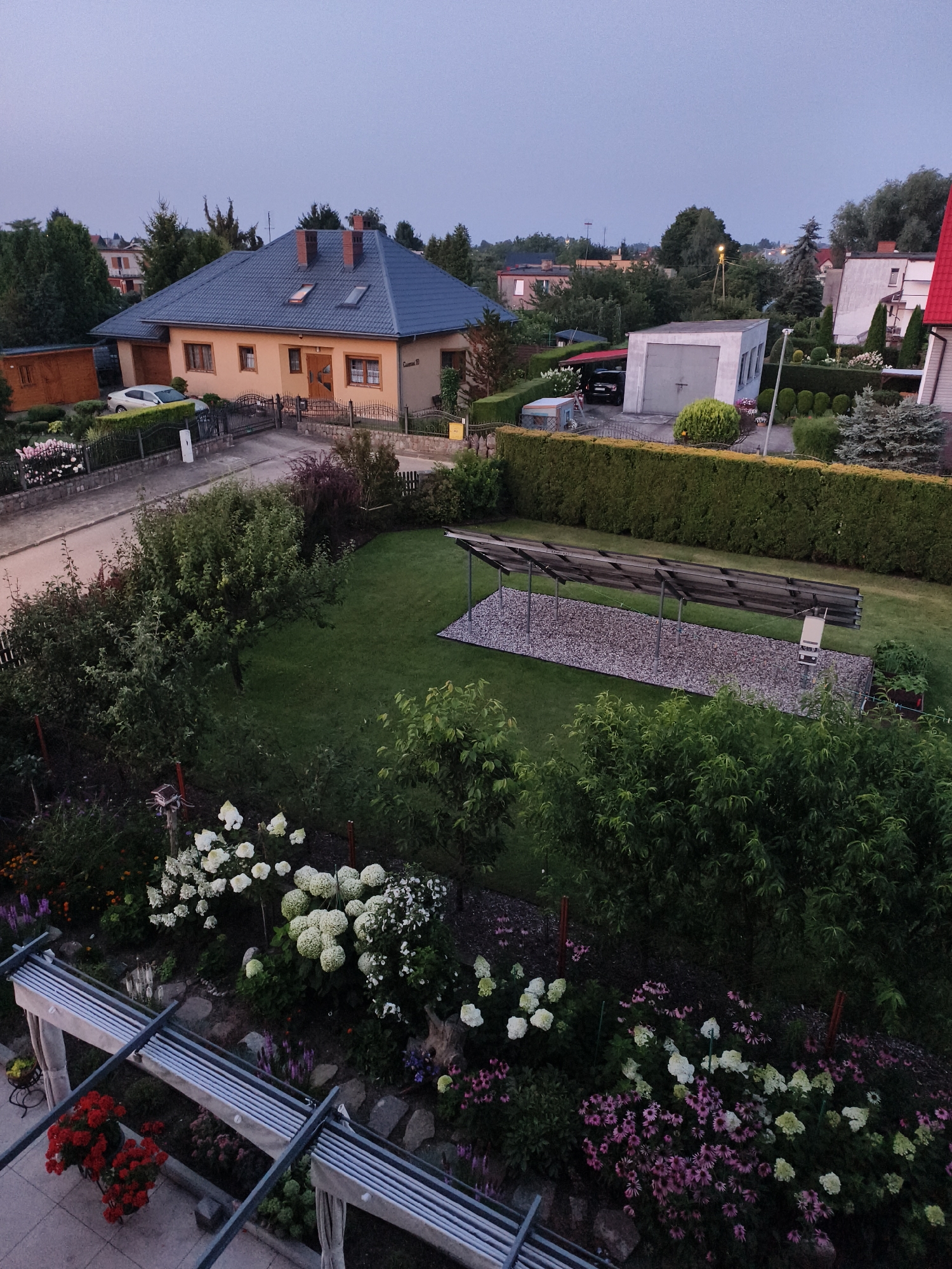 The image shows a residential area in Tczew, Poland, at dusk. In the foreground, a well-maintained garden features a variety of flowering plants, including hydrangeas and pink coneflowers, along with a modern pergola structure. Beyond the garden, a lawn is visible with a set of solar panels on a gravel base. Further back, houses with pitched roofs, a garage, and dense greenery are present, with a street and sidewalk running alongside the properties. The sky is overcast and dark, indicating the approach of night. No people are visible in the scene.