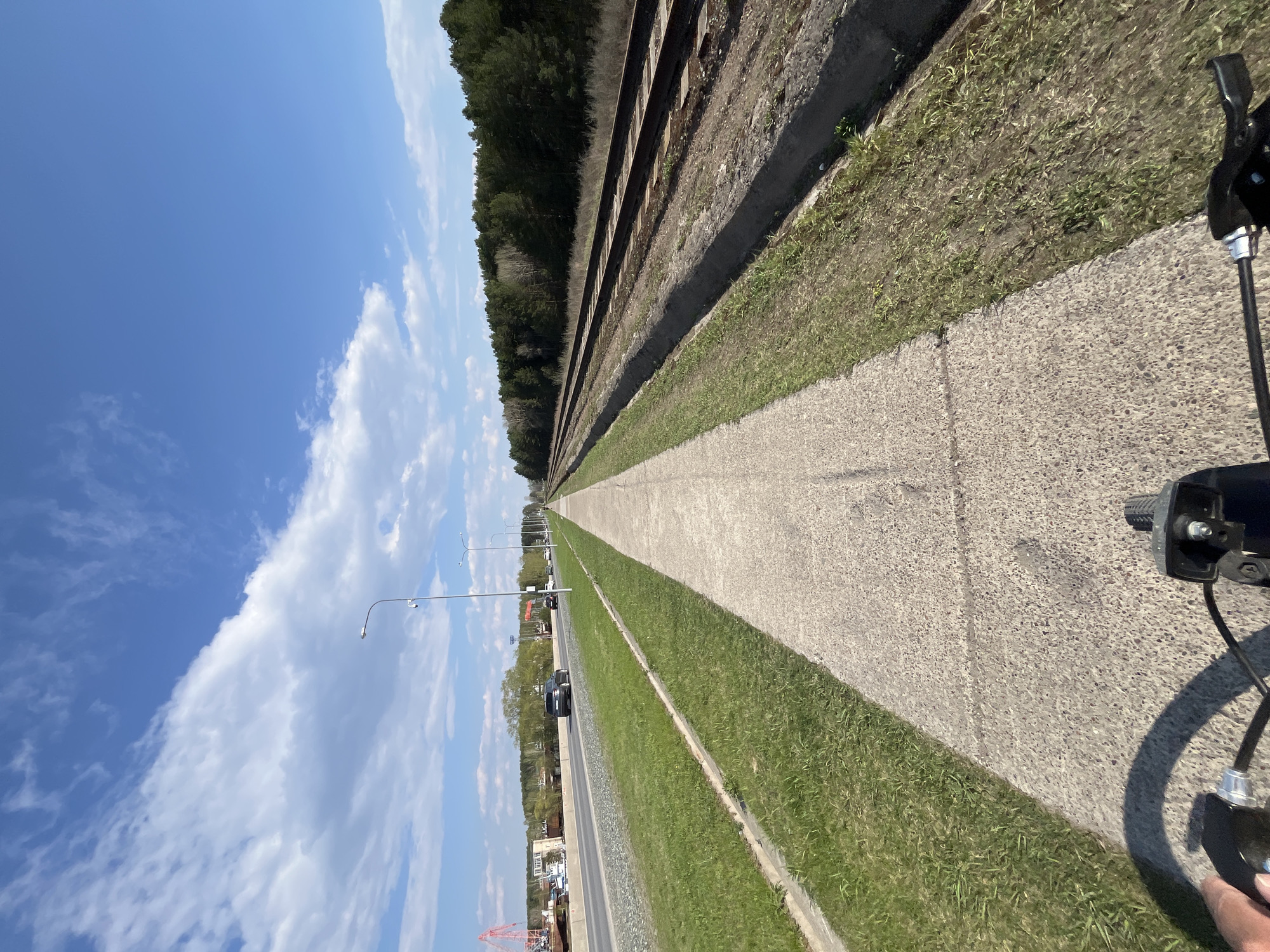 The image presents a perspective shot along a concrete path, flanked on one side by a grassy strip and then a railway track leading into a dense line of trees, and on the other side by another grassy strip bordering a road. Street lamps are sparsely spaced along the road. Several vehicles are visible on the road, including a dark-colored car. In the distance, construction activity with a crane is observed. The sky is blue with scattered clouds. Part of a scooter or bicycle is visible in the foreground. The location is identified as Chaikovsky, Russia.