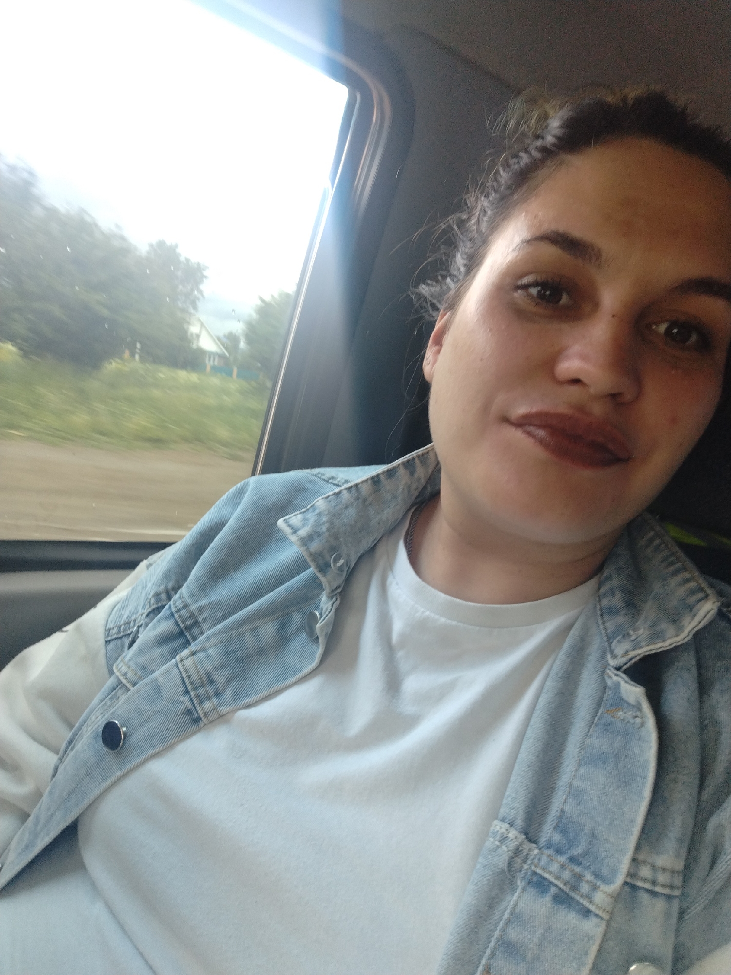A young woman is featured in a close-up, likely a selfie, taken inside a moving vehicle. She has dark hair styled in braids or twists, pulled back, and looks directly at the camera with a neutral expression, hinting at a slight smile. She is wearing a white t-shirt beneath a light blue denim jacket. Her lips are made up with a dark, glossy lipstick.
To her left, a car window shows a bright, partially overexposed rural landscape, with noticeable motion blur. Green fields and trees dominate the view, and a distant house with a light-colored facade and a blue fence is visible. The bright sky outside suggests it is daytime. The overall scene conveys travel in a rural or semi-rural area. No visible text is present in the image. The location context for this image is Фоки, Russia.