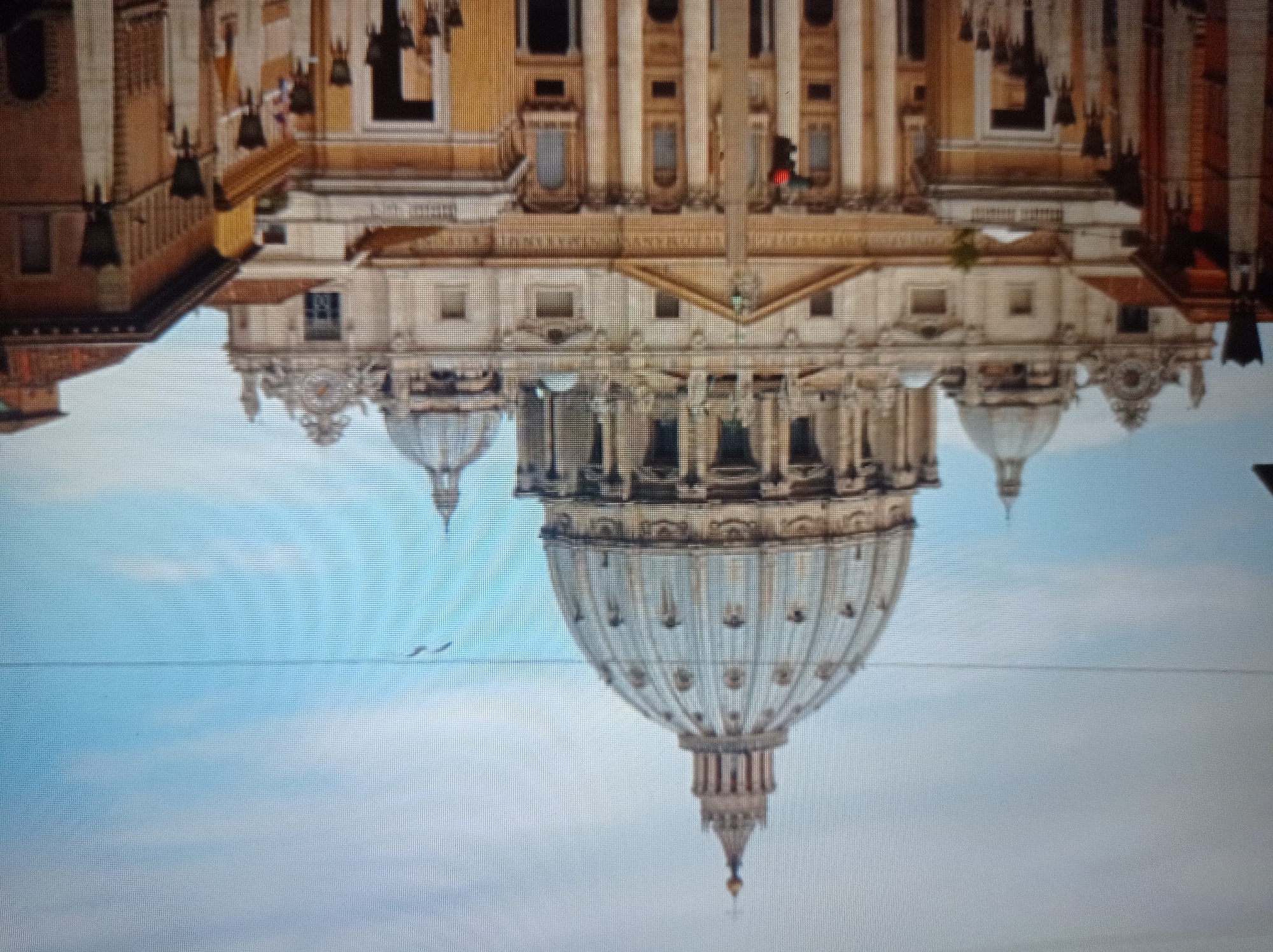 The image shows a wide-angle view of St. Peter's Basilica in Vatican City, Rome, Italy. The grand facade of the basilica dominates the frame, with its iconic dome prominently displayed against a cloudy sky. The surrounding buildings, characteristic of Roman architecture, frame the central structure. The scene depicts a clear day with a few birds flying in the distance. The overall impression is one of architectural grandeur and historical significance. There are no people visible in the image. The image is of an urban setting, likely a public square or street in front of the basilica. The lighting suggests it is daytime. The image is of good quality and shows sharp details of the architecture.