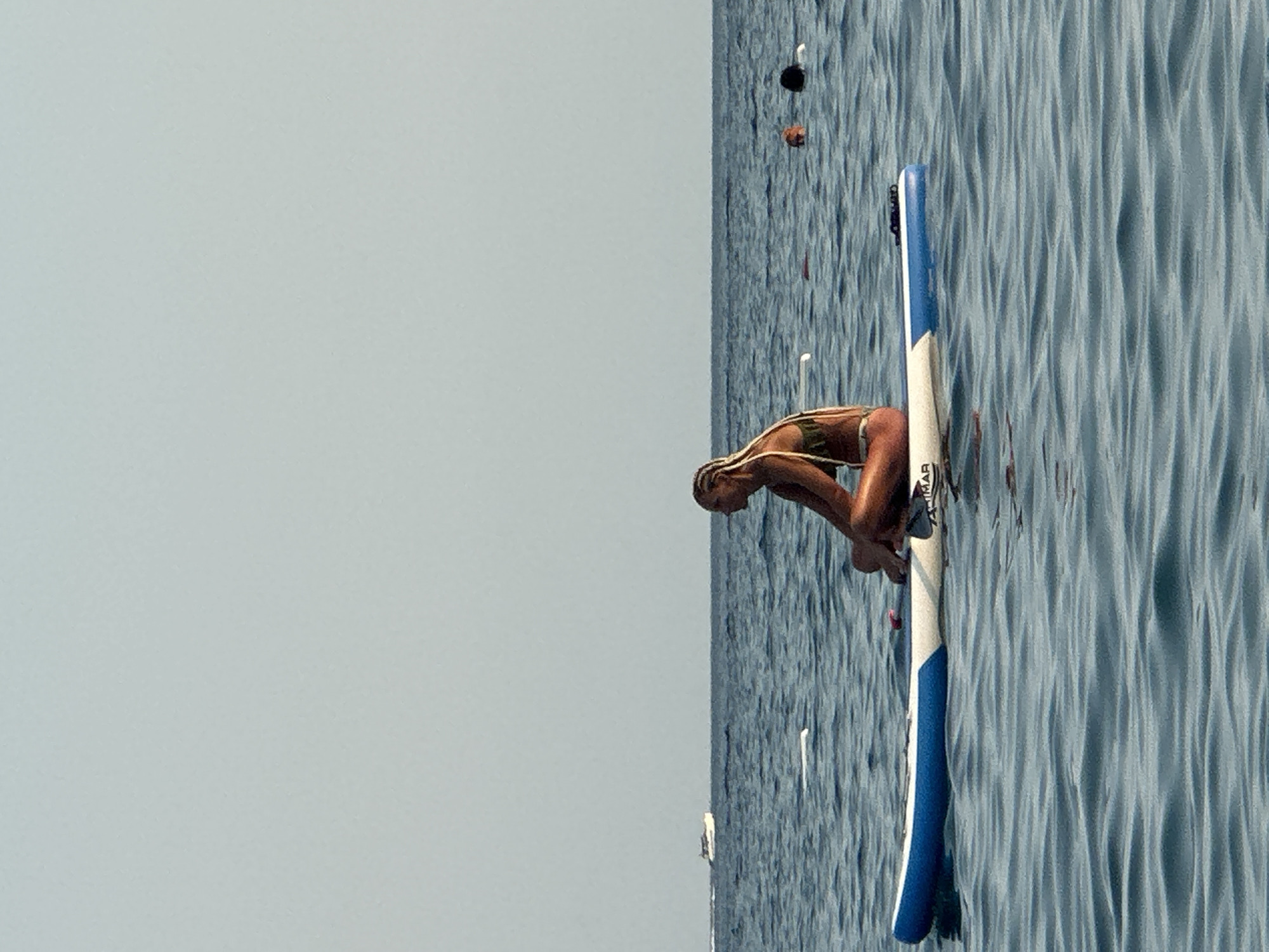 A person is sitting cross-legged on a blue and white paddleboard in the calm, blue water of the sea, likely off the coast of Boreti, Montenegro. The person, viewed in profile from the right, has blonde hair styled in braids and is wearing a patterned bikini. Their body is tanned. The paddleboard is angled slightly towards the left and has "ZIMAR" visible on its side. In the background, the sea stretches to the horizon under a clear, pale sky. Several other people are visible in the water further in the distance, appearing as heads. A white boat is also seen on the horizon. The water has small ripples.