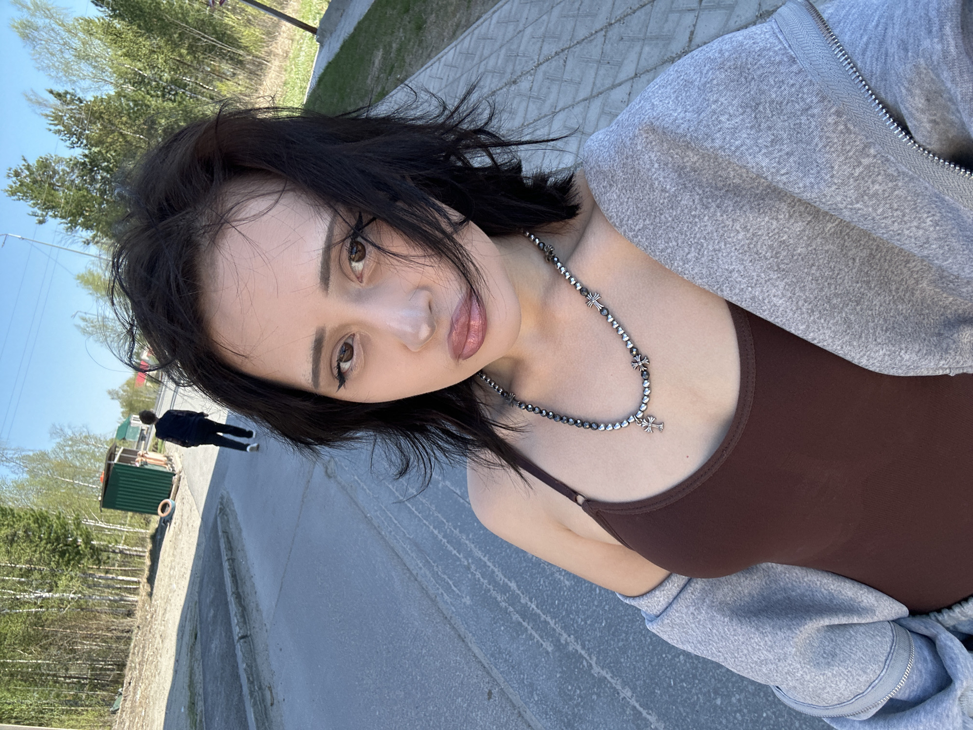 A woman is taking a selfie on a sidewalk in Высокий, Russia. She is wearing a brown tank top, a gray zip-up hoodie, and a necklace with cross pendants. She has dark hair and brown eyes. In the background, a person walks past a green kiosk. Trees and a clear blue sky are also visible. The sidewalk has patterned markings.