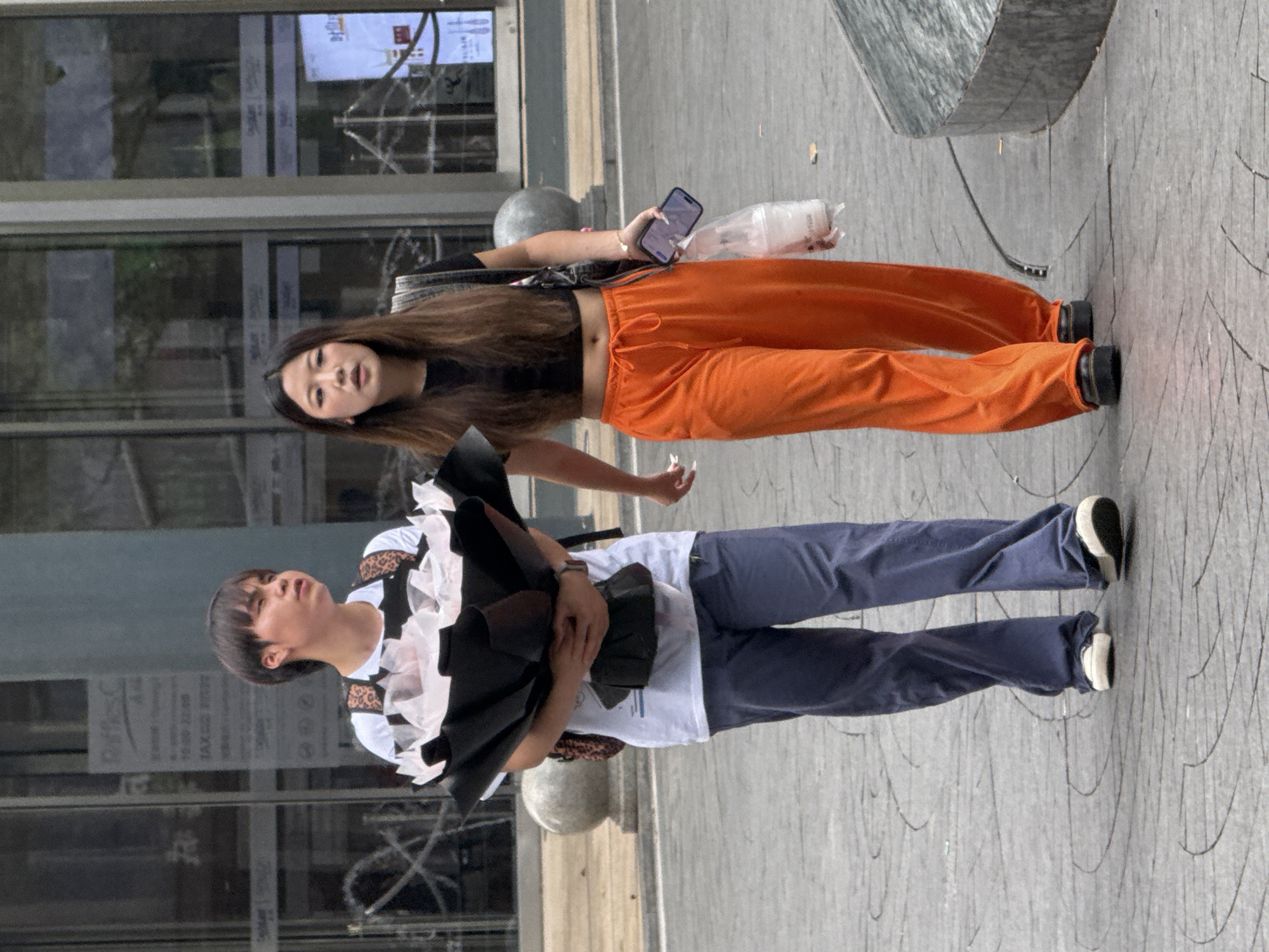 Two individuals are walking on a textured gray pavement in Yuzhong District, China. The person on the left, wearing a white t-shirt, blue pants, and white sneakers, is holding a large bouquet of flowers wrapped in black and pink material. This individual has dark hair and is looking to the right. To the right, a second person is walking, wearing a black crop top, bright orange wide-leg pants, and dark platform shoes. They are carrying a black backpack and holding a mobile phone in their right hand and a clear plastic bag with a drink in their left hand. The background shows a modern building with large glass windows and some signage. The scene appears to be outdoors during daylight.