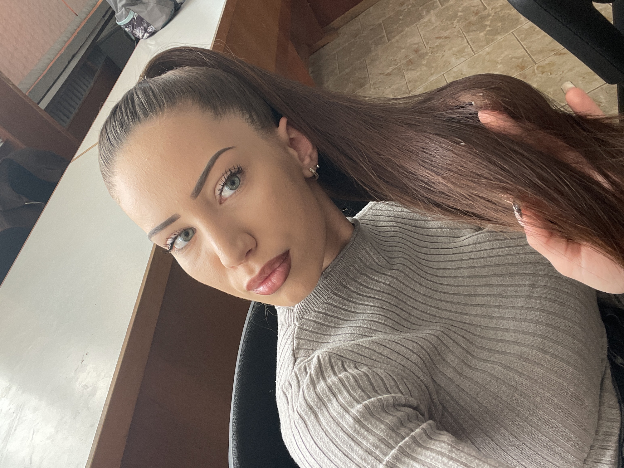 A young woman with light olive skin, high cheekbones, and green eyes is depicted in a close-up, high-angle shot. Her dark brown hair is pulled back into a high ponytail. She is wearing a light grey ribbed sweater. A small, simple hoop earring is visible in her left ear. The background includes part of a white table, a wooden cabinet, and a section of what appears to be a tiled floor. Part of a black chair is also visible, suggesting she is seated. The location is indeterminate from the given information.