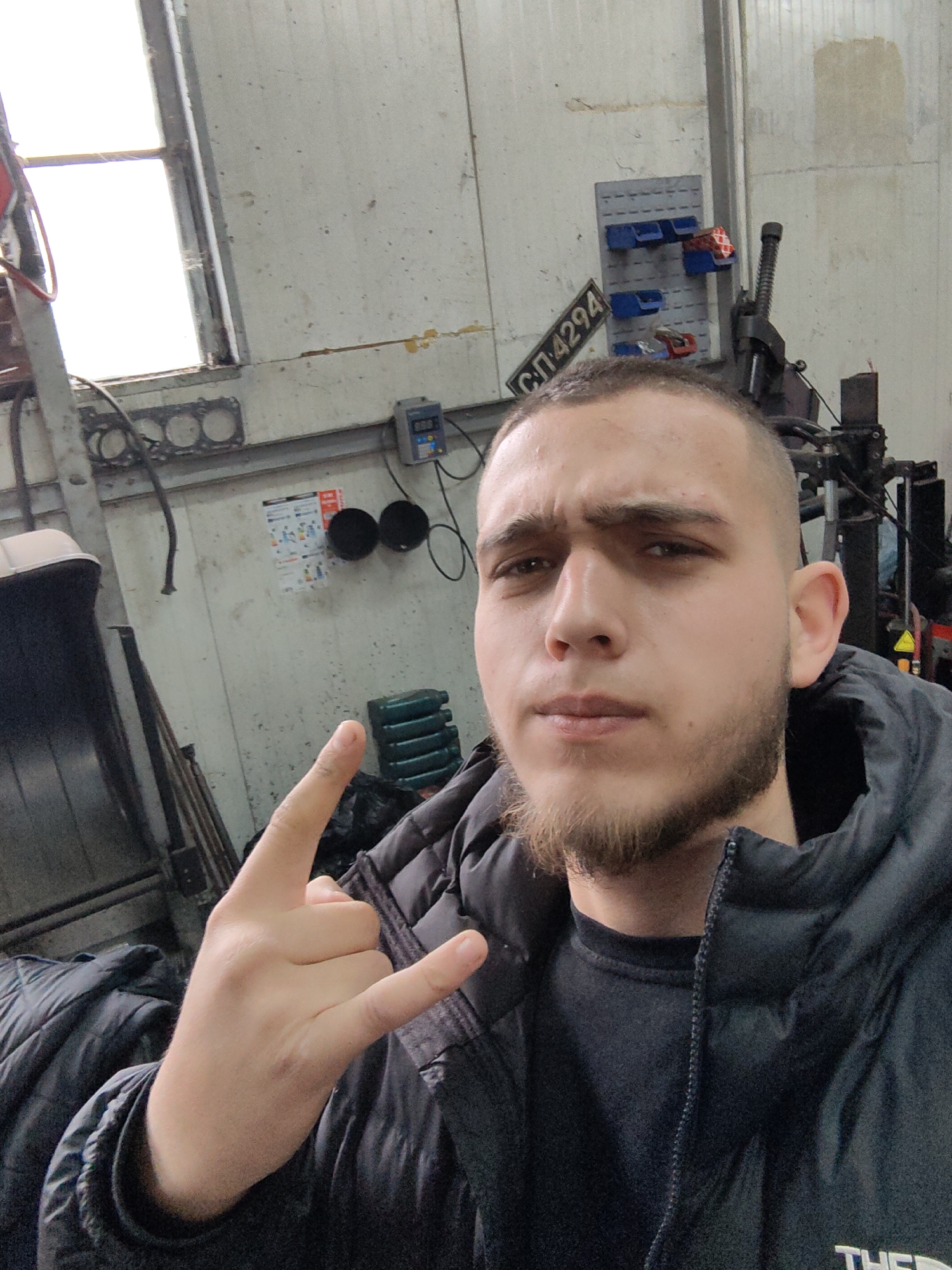 A man poses indoors, likely in a workshop or garage, based on the surroundings. The background includes a window letting in daylight, various tools, equipment, and storage solutions attached to the wall. A license plate reading "CN 4294" is visible on the wall behind him. The man is wearing a dark jacket and a t-shirt and is holding up the "rock on" hand gesture. The location is potentially in Sofia, Bulgaria, based on the license plate format commonly used in Bulgaria.