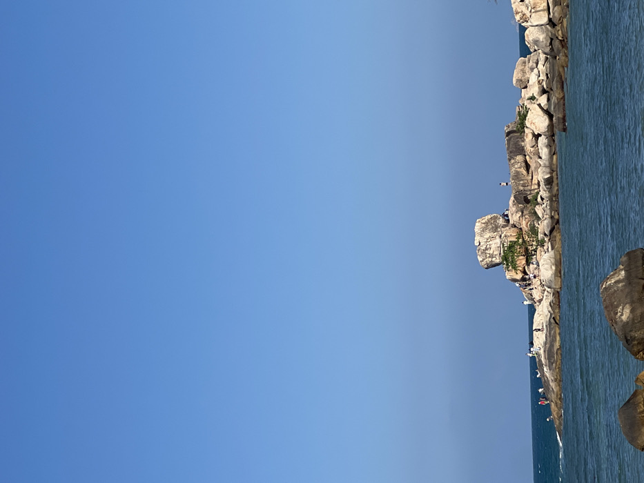 This image depicts the Hon Chong Promontory, a geological formation located in Nha Trang, Vietnam. The scene is dominated by a clear, unblemished light blue sky occupying approximately two-thirds of the vertical frame. Below, a prominent cluster of large, irregularly shaped granite boulders forms a rocky outcrop extending into the sea. A notable feature within this formation is a large, rounded boulder balanced on top of other rocks towards the left side of the main outcrop. Sparse green vegetation is visible growing between some of the rocks.
Approximately two dozen individuals are observed on the rock formations, distributed across various ledges and peaks. They appear to be tourists or visitors, standing, walking, and observing the surroundings. Their postures suggest casual exploration. The sea water, visible in the foreground and surrounding the rocks, is a deep blue-green, showing minor surface ripples and small white waves crashing gently against the base of the rocks on the far left. Several dark, partially submerged boulders are present in the immediate foreground. The lighting is bright and even, indicative of clear weather and strong daylight, casting minimal shadows.