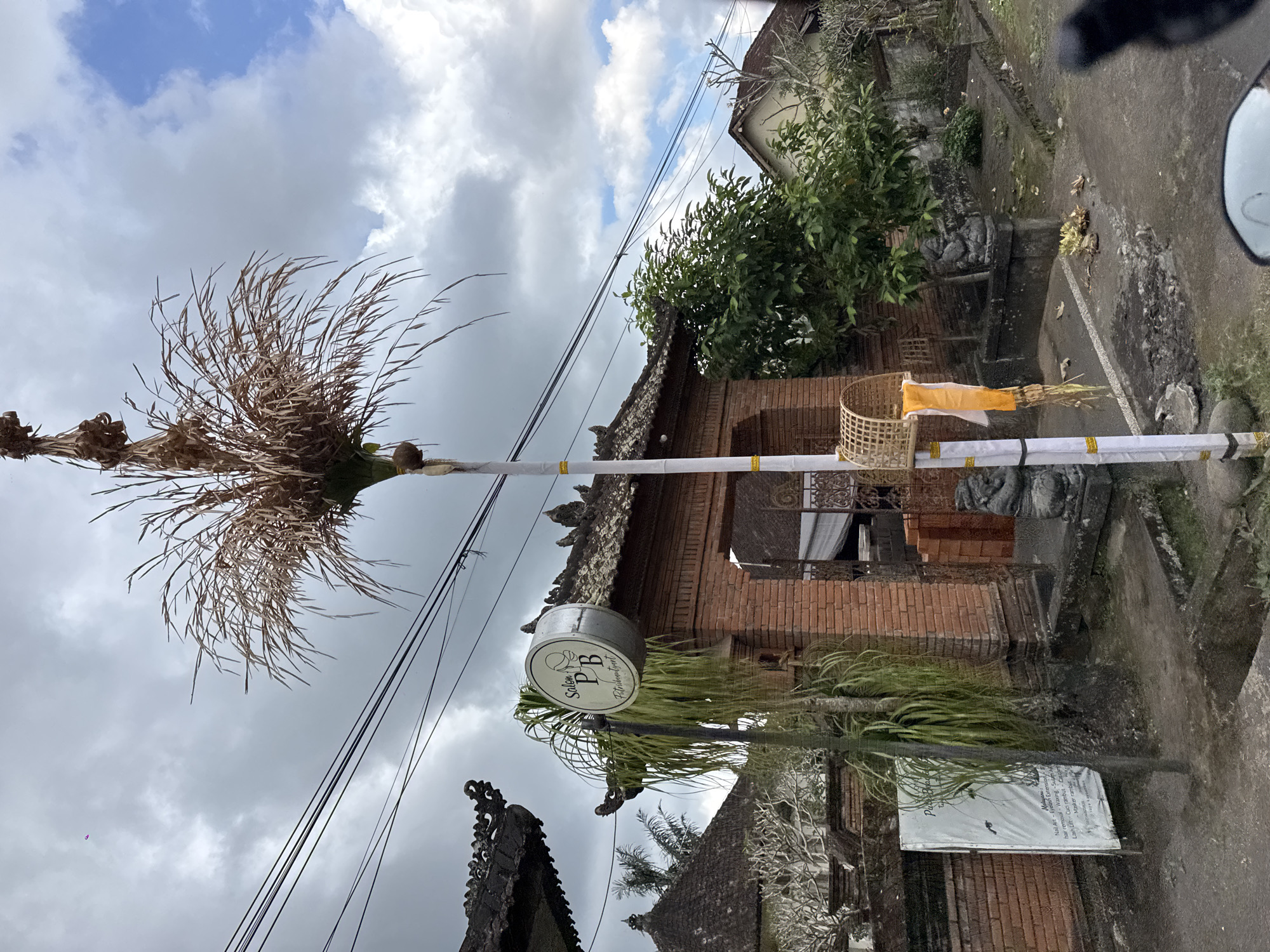 This vertical image captures an outdoor scene in Susut, Indonesia, likely on a cloudy day. The foreground is dominated by a tall, decorative bamboo pole adorned with a woven basket containing a yellow cloth and some dried vegetation. A cylindrical sign hangs from the pole, displaying text including "Salon PB" and "Potribeautyart" along with other services like "Nail Art," "Eyelash Extension," and "hair removal."
Behind the pole, a traditional Balinese brick building with intricate roof details is visible. Lush green trees and plants surround the building, indicating a tropical environment. The sky is filled with heavy, grey clouds, suggesting an overcast or potentially rainy day. Power lines crisscross the upper portion of the frame, indicating the presence of modern infrastructure within the traditional setting. The overall impression is one of a local business or establishment set against a backdrop of Balinese architecture and vegetation.