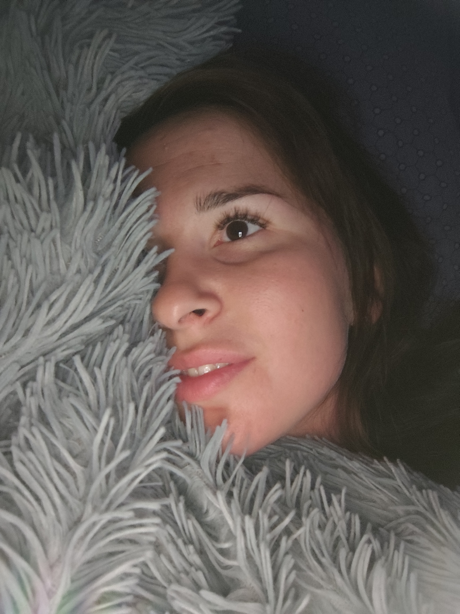 A close-up, high-angle indoor shot shows a woman partially covered by a fluffy gray blanket. Her face is turned to the right, with her right eye and part of her left eye visible, along with her nose and mouth. She has dark hair and fair skin. The background is dark and out of focus, with a subtle textured pattern. No specific location in Фоки, Russia is identifiable.