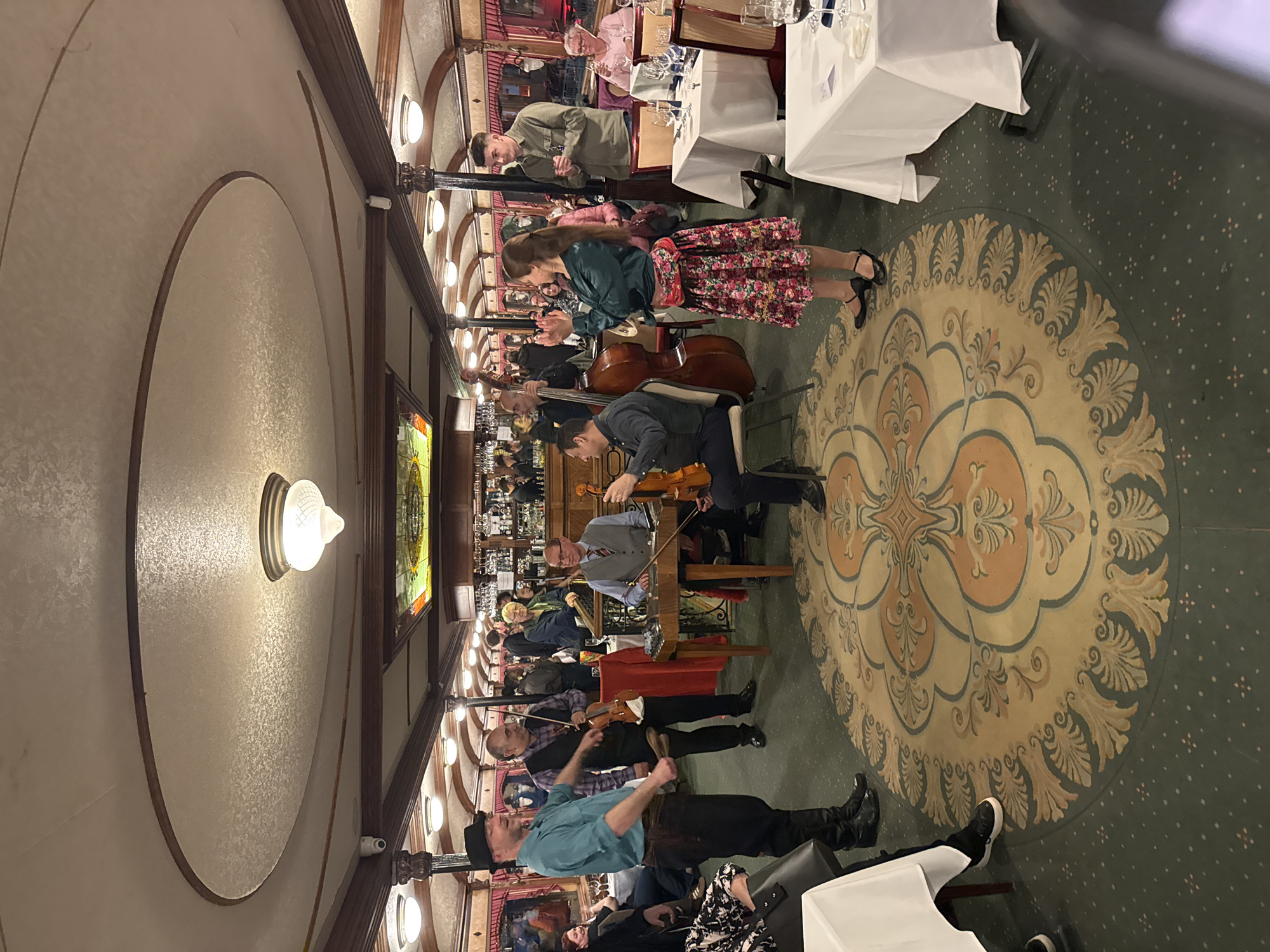 This image depicts an indoor scene within what appears to be an elegant restaurant or dining hall, where a live musical performance is taking place for patrons. The setting is richly decorated with a classic aesthetic.
In the mid-ground, a musical ensemble is actively performing. Visible instruments include a double bass, at least two violins, a cello, and a hammered dulcimer or similar stringed instrument. The musicians are dressed in smart casual attire, engaged in their performance. Several people are observing the performance, including a woman in a floral skirt and dark top standing near the musicians.
The background shows multiple dining tables, each covered with a white tablecloth and set with glasses and dinnerware. Patrons are seated at these tables, some appearing to be dining, while others are observing the band. Behind the dining area, a well-stocked bar is visible, filled with bottles and glasses.
The floor is covered with a patterned carpet, featuring a large, ornate circular motif in shades of cream, gold, and light blue-green in the center, surrounded by a dark green carpet with smaller speckled patterns. The ceiling is arched and features dark wooden beams and decorative elements. Warm, artificial light emanates from multiple wall sconces and a large, central light fixture, indicating the event is likely taking place during the evening or night. Architectural details include decorative pillars and a stained-glass window panel.
The scene captures an atmosphere of sophisticated entertainment and dining. No visible text is discernible in the image.