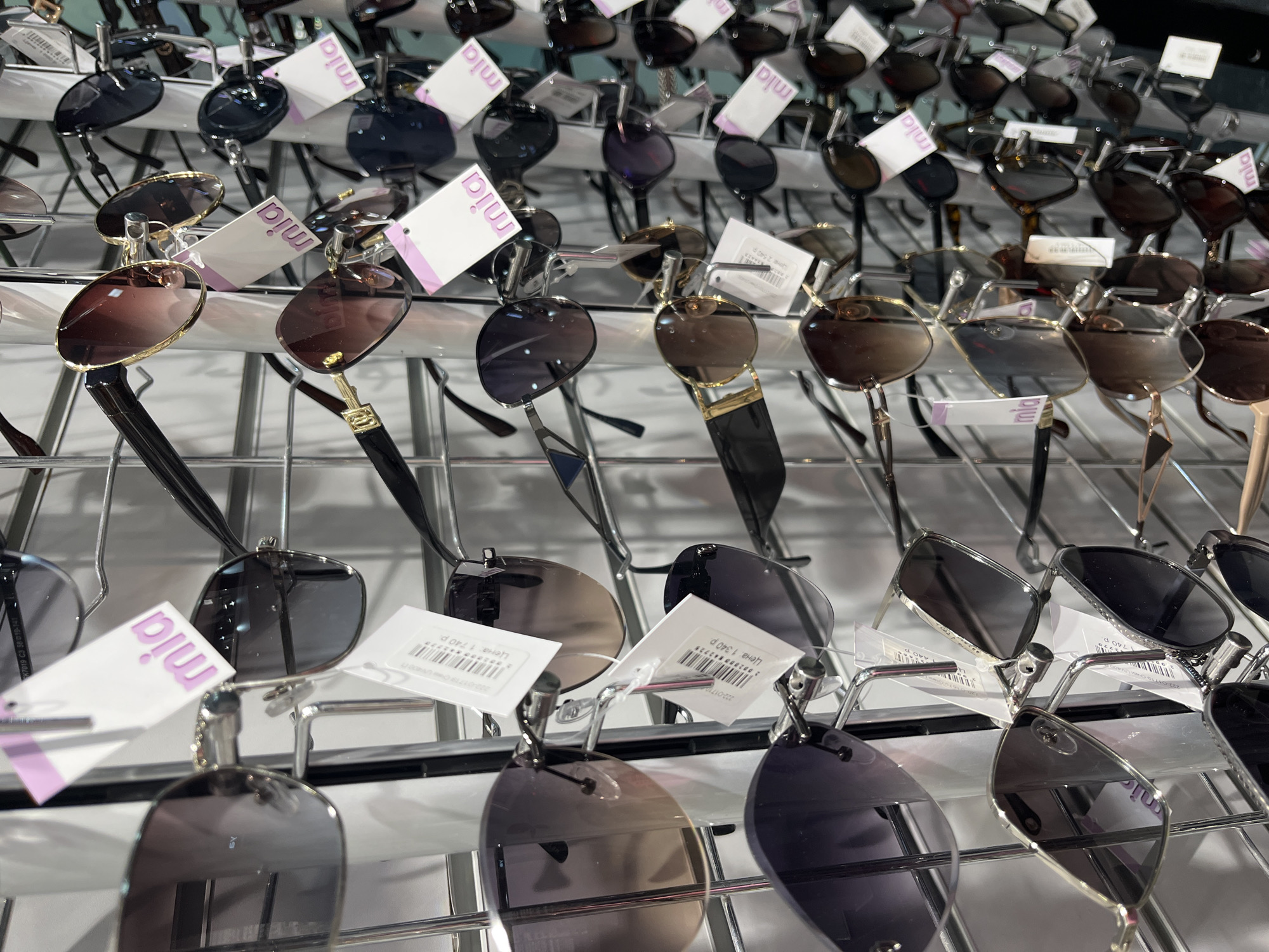 The image shows a display of sunglasses, likely in a retail setting. The sunglasses are hanging on a wire rack. Many of the sunglasses have tags attached with the brand name "mia" and price tags in Russian. Price tags visible show amounts of 1340, 1740, and 2540 rubles. A tag on a pair of sunglasses shows the frame specs to be K7019 C3 58 019-141. The scene appears to be indoors, possibly in a shop or market in Izhevsk, Russia, based on the Russian language on the price tags.