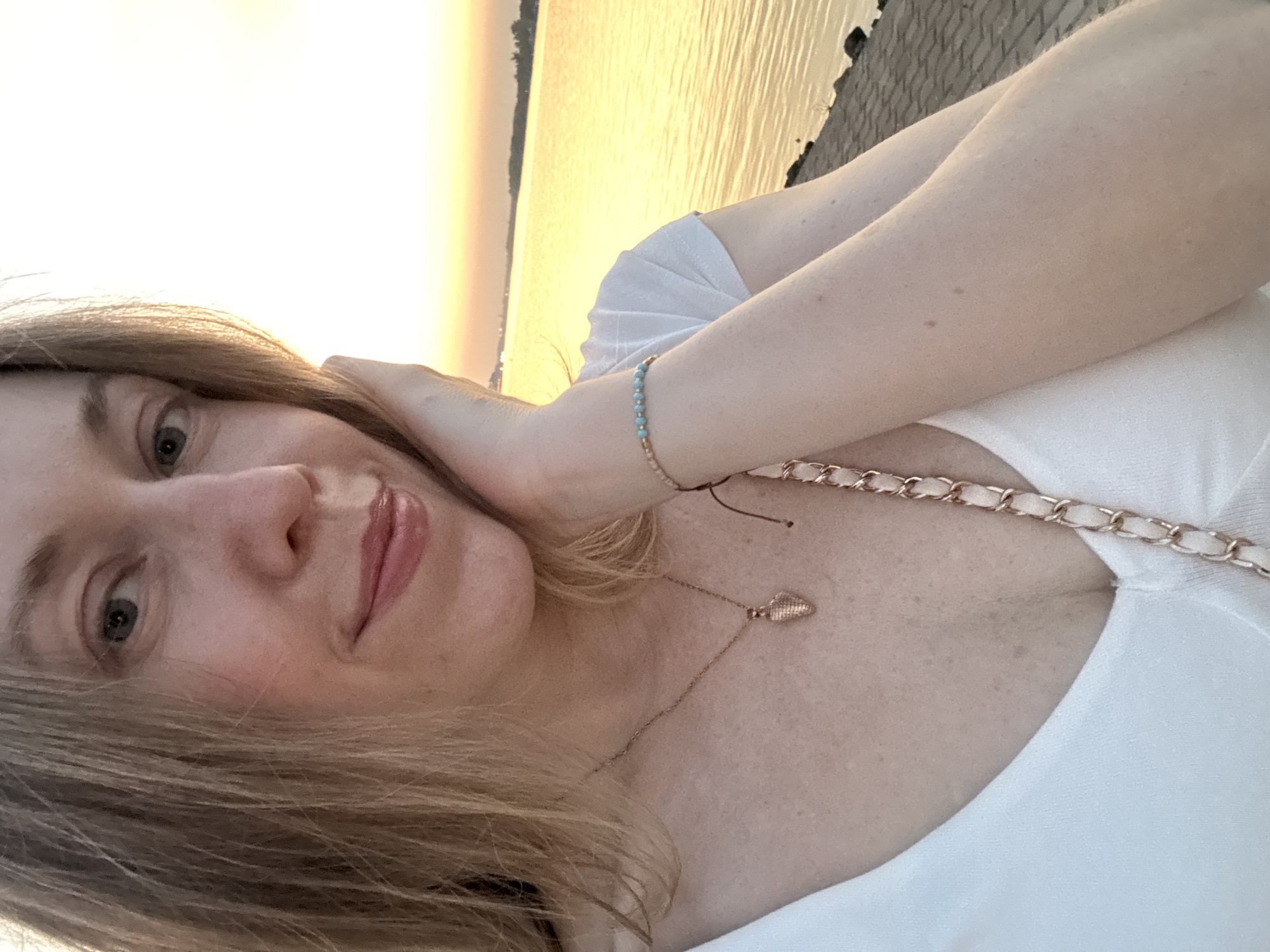 A woman with blonde hair takes a selfie near a body of water at sunset. She is wearing a white t-shirt, a necklace with a heart-shaped pendant, and a bracelet. The water appears to be a river, reflecting the warm hues of the setting sun. The background shows a distant shoreline with buildings and trees. The foreground consists of a paved walkway along the water's edge. The image is taken from a medium close-up angle, focusing on the woman's face and upper body. This scene is likely from the waterfront of Belgrade, Serbia, given the characteristic urban riverside landscape and the warm, golden light typical of sunsets over the Sava or Danube rivers.