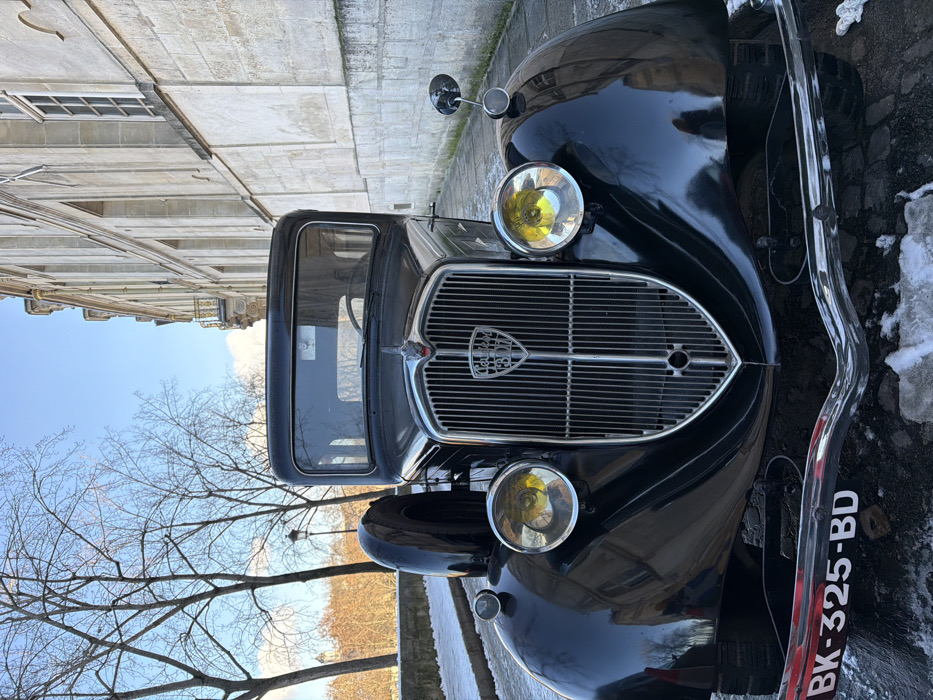 A black vintage Peugeot automobile, likely from the 1930s, featuring a vertical chrome grille with a lion emblem, large circular yellow-tinted headlights, and a fender-mounted spare tire, is centrally positioned. Its front license plate displays "BK-325-BD". The car is parked on a paved surface partially covered with melting snow and slush. To the vehicle's right stands a tall, multi-story stone building exhibiting classical Parisian architecture, including arched windows and a small golden balcony on an upper floor. To the left, a low stone wall supports a line of bare deciduous trees. Beyond the trees, in the mid-distance, the distinctive dome of the Panthéon is visible under a clear blue sky with scattered clouds. The scene is set on a street in the Latin Quarter of Paris, France, likely on or near Rue des Écoles. No individuals are present.