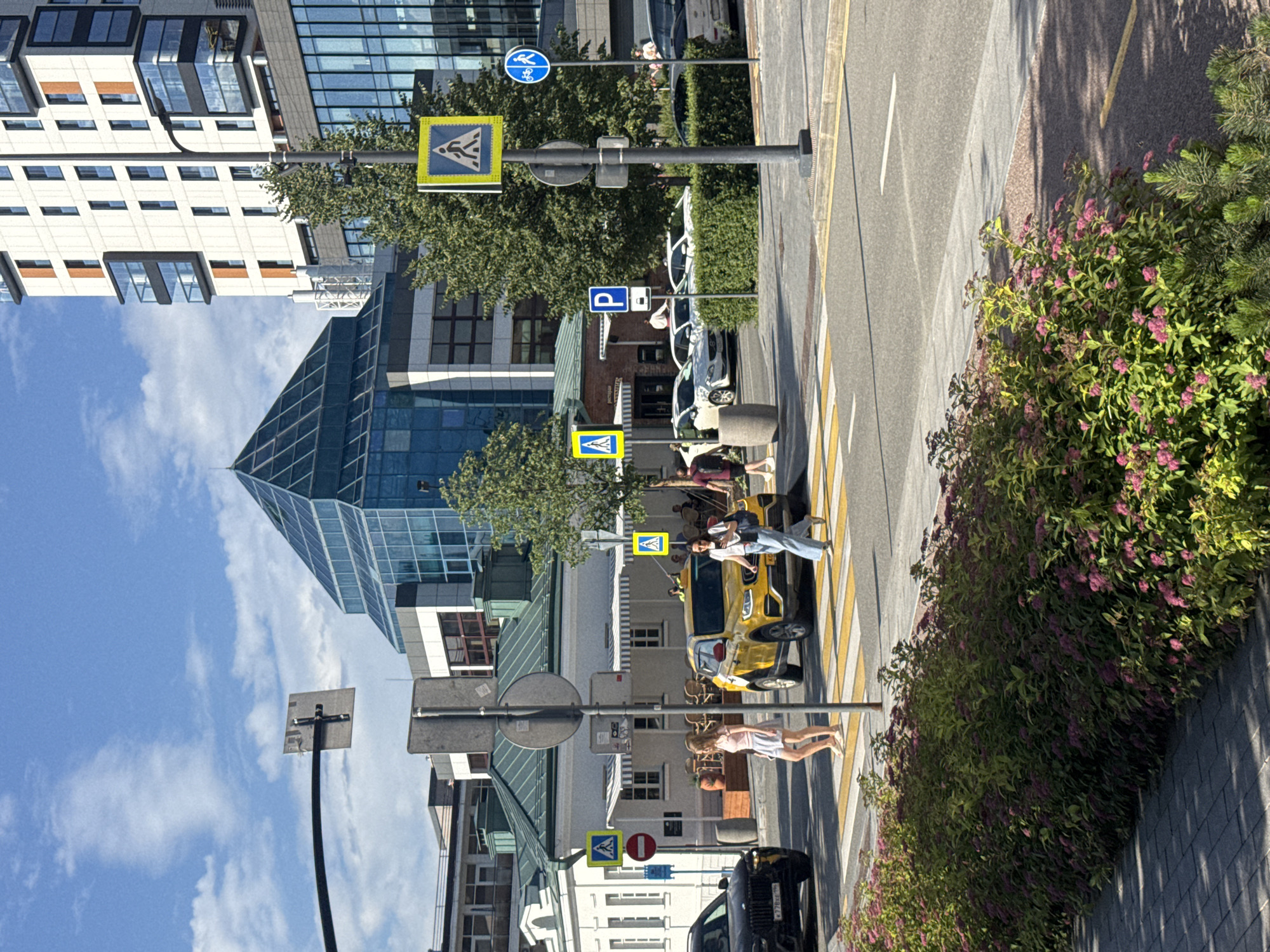 This media file captures an outdoor scene on a sunny day, likely in Yekaterinburg, Russia. The image is oriented vertically, showing a street with a crosswalk in the foreground. Several people are visible, some crossing the street and others standing near it.
In the mid-ground, a yellow car is parked on the side of the road. There are several signs visible, including pedestrian crossing signs, a parking sign, and a traffic sign with a red circle and a diagonal bar, indicating "no entry" or a similar prohibition. Buildings with modern architecture, featuring glass facades and balconies, line the street. Lush green trees and bushes with small pink flowers add to the urban landscape.
One person, a woman in a light pink dress, is walking across the crosswalk. Another group of people, two women and one man, are near the yellow car. The lighting suggests it is daytime, possibly late morning or early afternoon, with clear blue skies and scattered white clouds. The overall atmosphere appears casual and everyday, with no overt signs of distress or excitement. The image provides a snapshot of urban life with a focus on pedestrian activity and street elements.