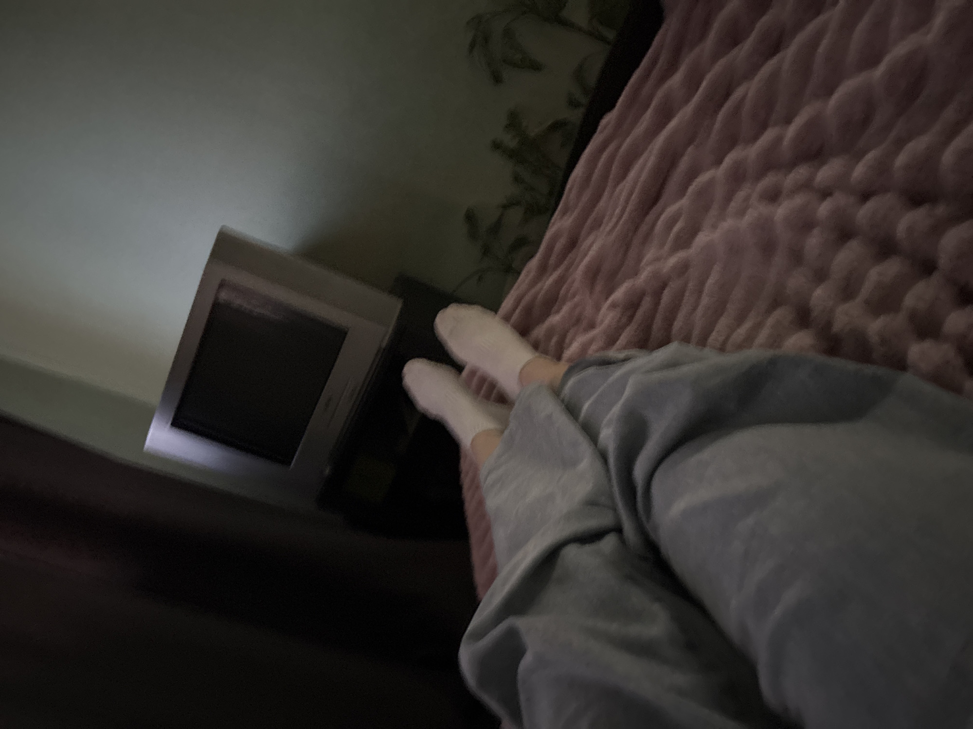 The image depicts a first-person perspective of someone lying in bed, looking towards a television. The room is dimly lit, with a dark curtain on the left and a pale green wall on the right. A vintage television set sits on a black stand. The person is wearing grey sweatpants and white socks. The bed is covered with a thick, pink knitted blanket. The overall impression is one of a relaxed, indoor setting, possibly a bedroom. The location is unknown, Russia.