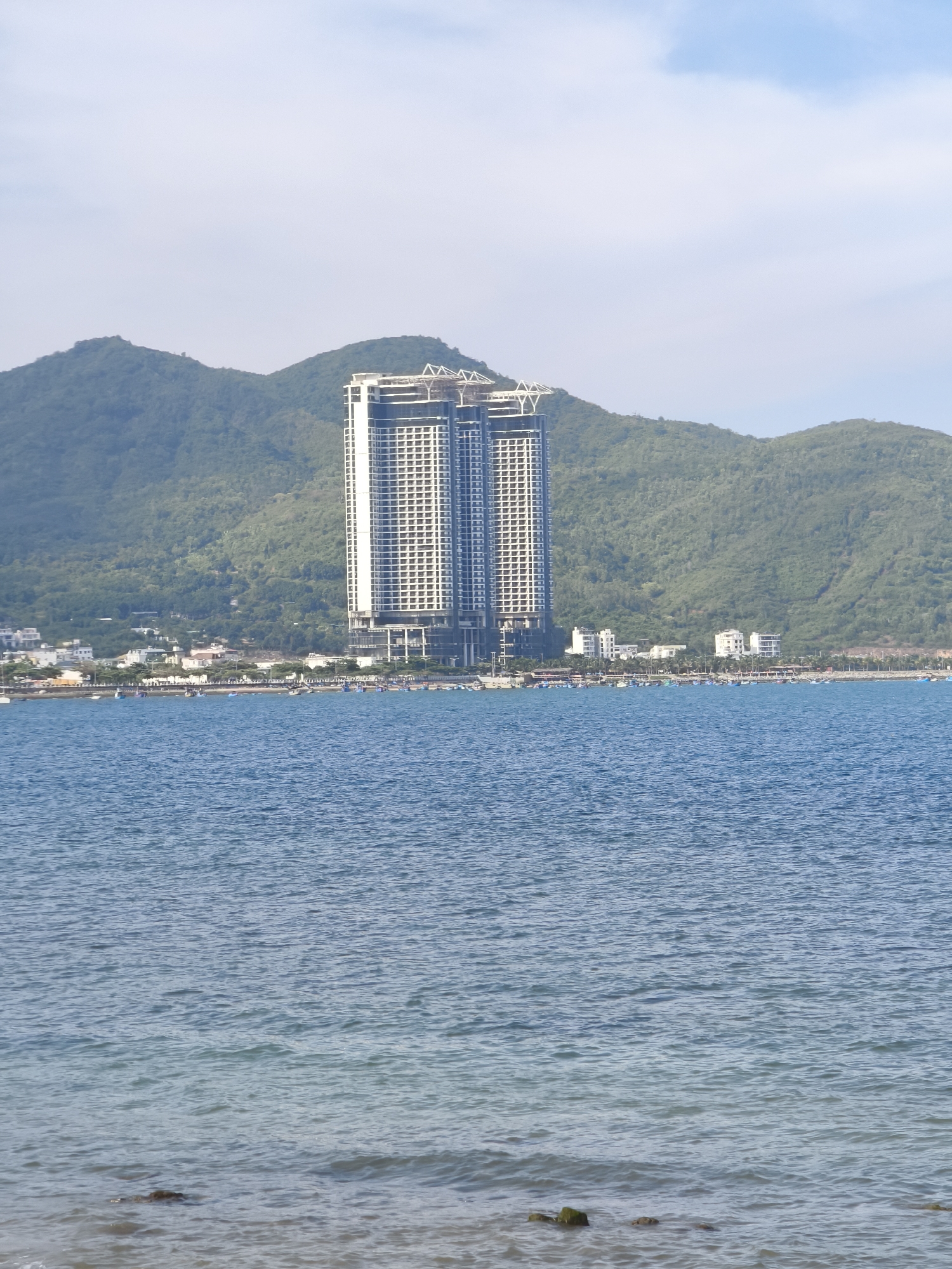 The image shows a coastal scene in Nha Trang, Vietnam, dominated by the ocean in the foreground and a densely forested mountain range in the background. A prominent, modern, twin-towered skyscraper stands in the middle ground, centrally located against the mountainside. Below the skyscraper, a strip of buildings forming a cityscape runs along the coast, interspersed with palm trees. A line of small boats is visible on the water in front of the cityscape. The ocean water is a calm blue, with gentle waves lapping at a rocky shoreline in the immediate foreground. The sky above is pale blue with scattered white clouds.
