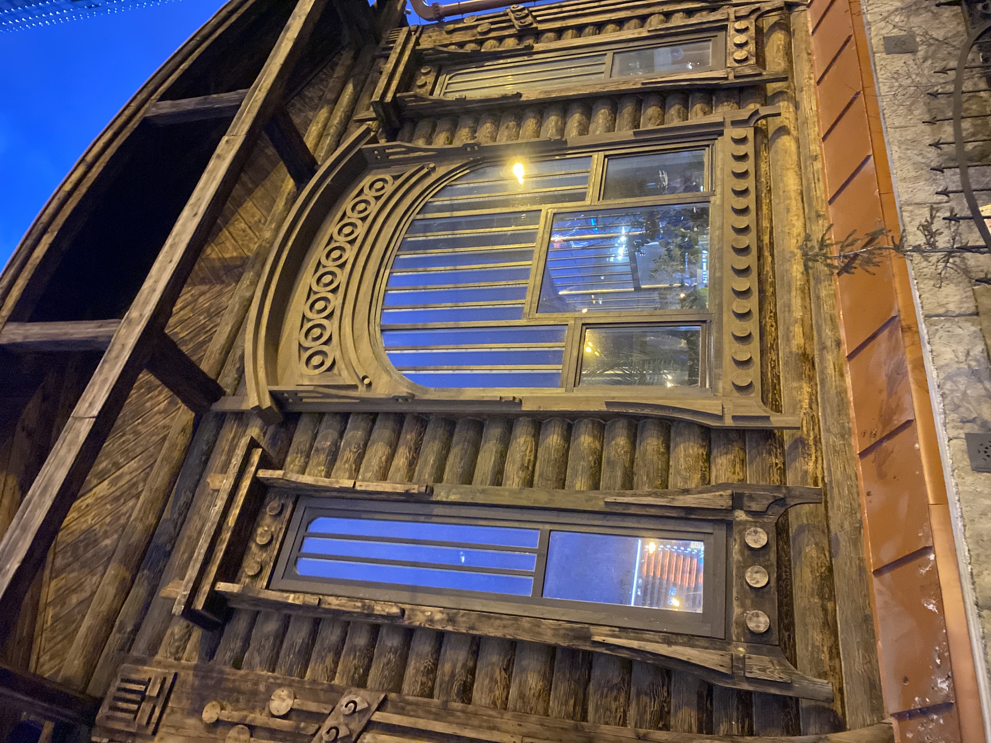 This media file captures a unique, intricately designed building facade, likely at dusk or night, under a deep blue sky. The building exhibits a distinct, rustic architectural style, primarily constructed from dark, weathered wood.
The dominant feature is a large, semicircular arched window, framed by ornate wooden carvings depicting a series of interconnected circles. This window, along with others, reflects interior lights and elements, including hints of greenery. Above this prominent window, and across the facade, are smaller rectangular windows with multiple panes, some showing internal lights or reflections of outside light sources. The wood on the building's left side is curved and planked, reminiscent of a ship's hull, while other sections feature vertically arranged wooden logs and carved decorative elements like crescent shapes and geometric patterns.
To the right, an adjacent part of the building or a neighboring structure features a metallic facade, possibly copper or rusted steel, characterized by diagonal grooves. This section is next to a lighter-colored wall, likely stone or concrete, from which some sparse, dark foliage or branches protrude.
The overall scene suggests a creative and unconventional architectural design, possibly for a commercial establishment like a restaurant or shop, given its detailed and inviting appearance under artificial lighting. There is no visible text. The time of day is clearly evening or night due to the dark sky and artificial illumination. The location is Yekaterinburg, Russia.