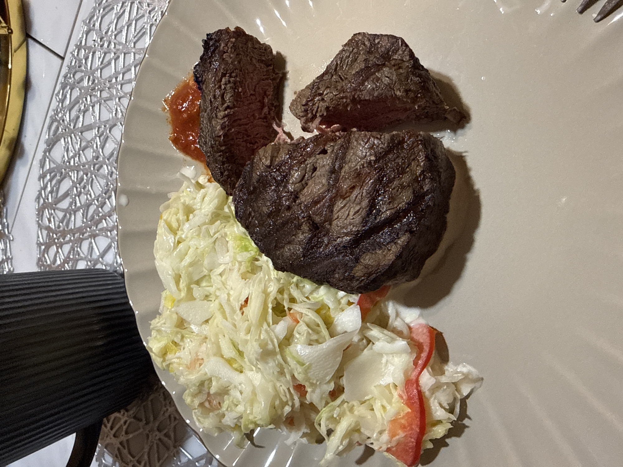 An overhead close-up shot displays a meal served on a light gray dinner plate. The main subjects are several pieces of cooked meat, likely steak, and a large serving of coleslaw.
The meat pieces, dark brown with grill marks on the exterior, are cut to reveal a brownish-pink, medium-well interior. A small quantity of reddish sauce or gravy is visible beneath one of the meat portions. The accompanying coleslaw is composed primarily of shredded white cabbage, mixed with thin strips of red bell pepper and other light-colored ingredients, all coated in a creamy dressing.
The plate rests on a table, part of which is covered by a metallic-looking placemat featuring an intricate, woven silver or light gray pattern, visible along the left and bottom edges of the frame. A dark, ribbed object, possibly a piece of furniture, is partially visible on the far left. A silver fork is seen in the upper right corner of the plate. The indoor lighting suggests an evening setting for this meal in Khimki, Russia. No text is visible in the image.