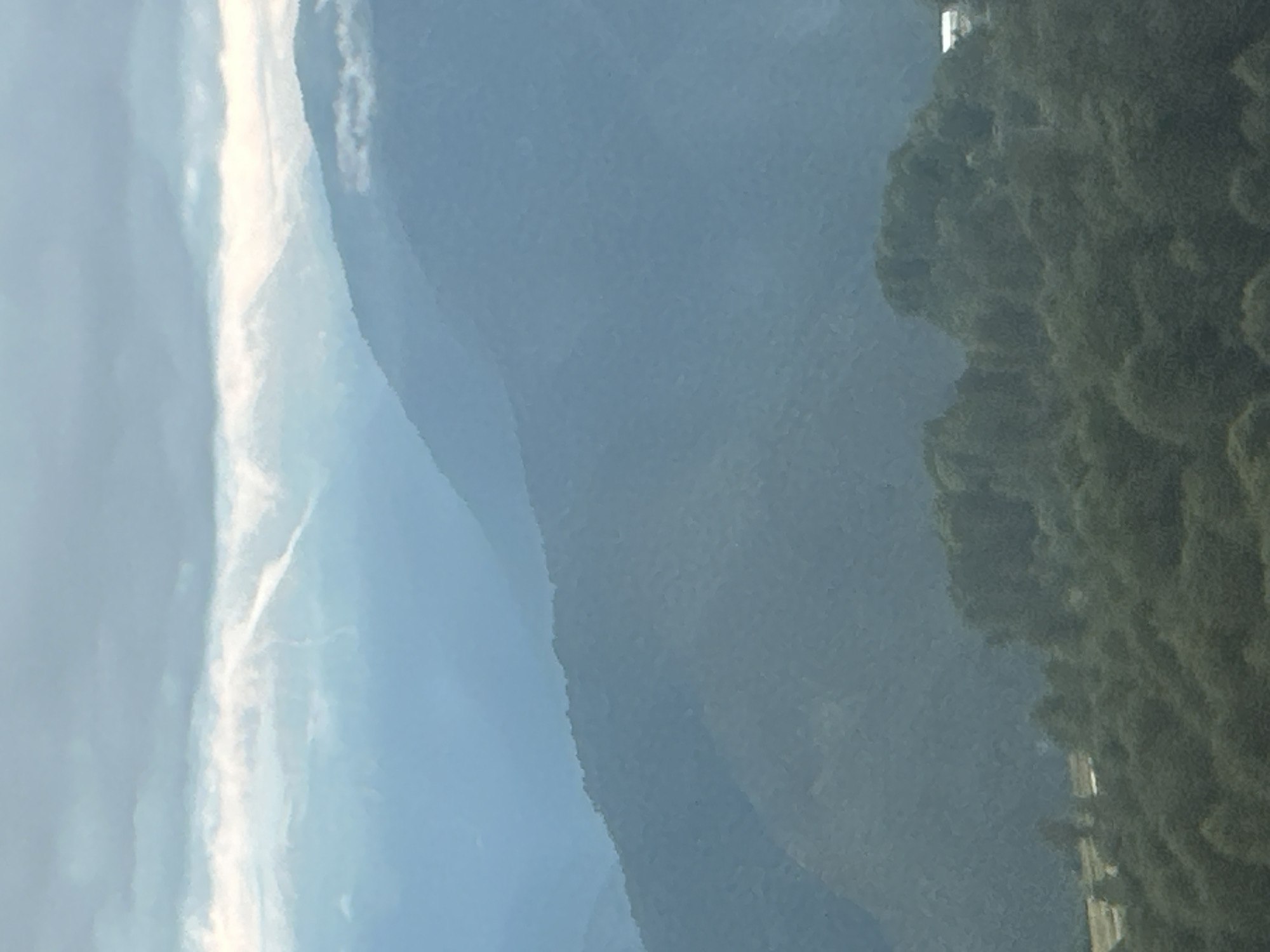 The image depicts a hazy, mountainous landscape with several layers of peaks receding into the distance. The foreground is dominated by dark green, dense foliage, likely trees. A small, white building is visible nestled within the trees on the right side of the frame. The sky is overcast with light grey clouds. The overall impression is one of a cool, damp, or foggy mountain environment. The scene is consistent with the topography of Estosadok, Russia, a resort town known for its mountainous terrain.