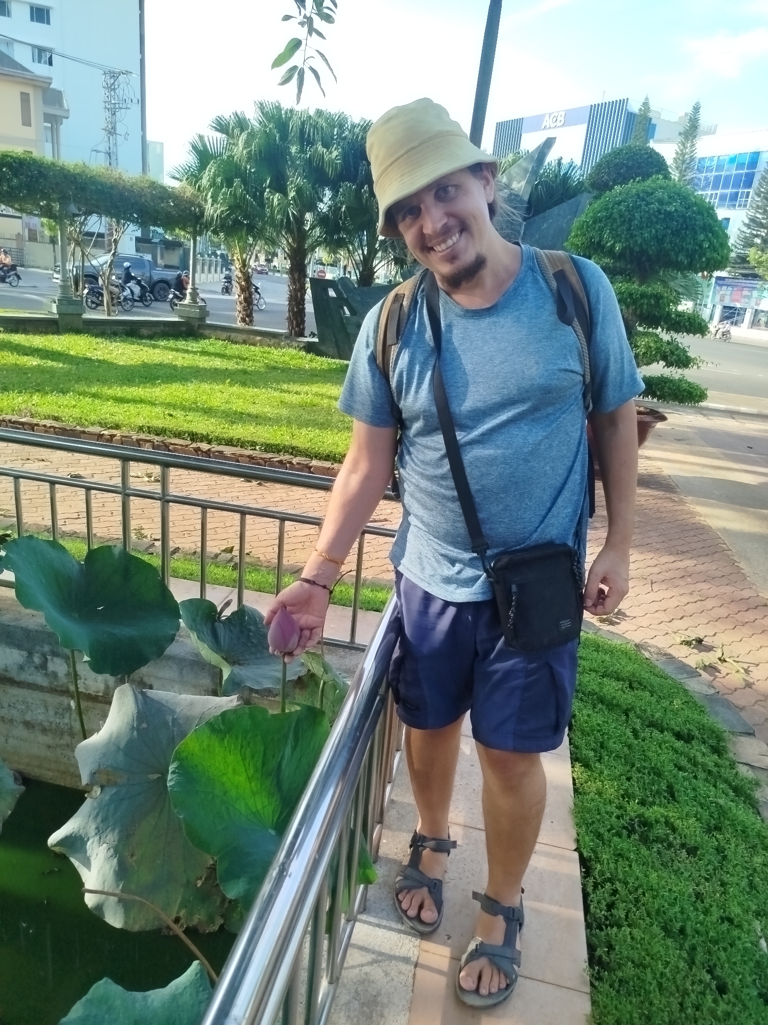 A man stands in a park in Pleiku Ward, Vietnam. He wears a khaki bucket hat, blue t-shirt, navy shorts, and grey sandals. A black messenger bag hangs across his torso, and he has a backpack. He stands next to a metal railing, holding a closed pink lotus flower over a pool of water with large green lotus pads. A brick-like walkway is to his right, bordered by a green ground cover. In the background are tropical trees, a street with motorbikes, and a modern building with the letters "ACB" visible. The day is sunny.