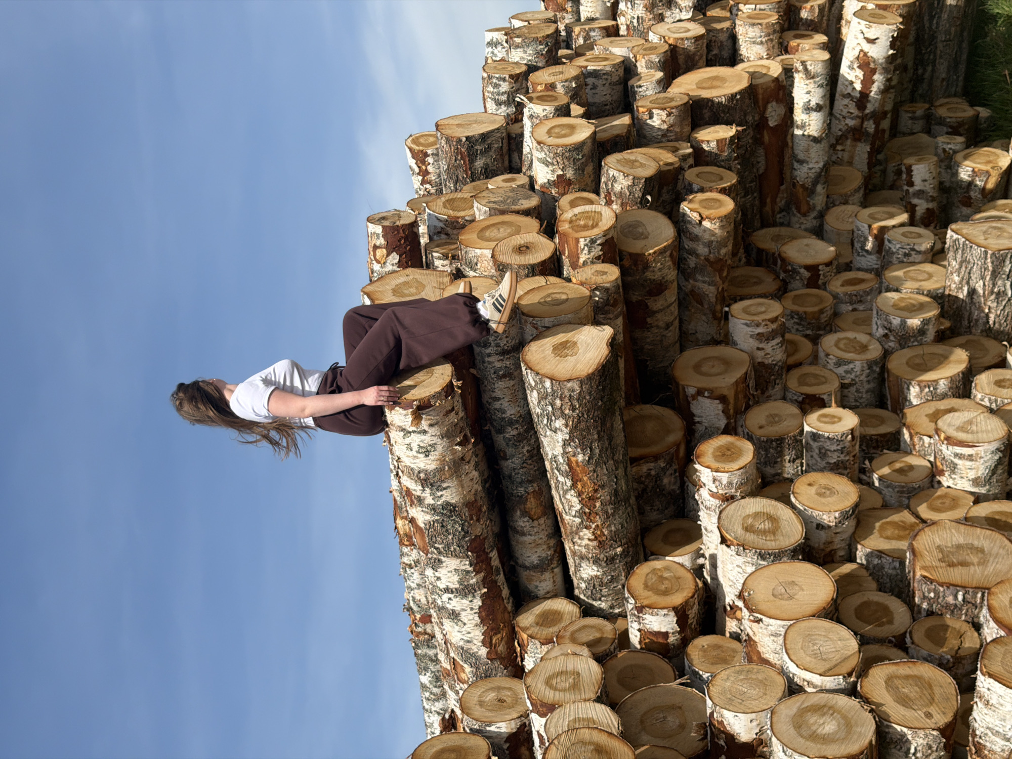 The image depicts a young woman lying on a stack of birch logs against a blue sky. The logs are neatly stacked, showing their cut ends and distinctive white bark. The woman is wearing a white t-shirt and brown trousers, with one foot visible wearing a sneaker. The perspective is tilted, making the sky appear to be on the side. The location is identified as Ural'skoe, Russia.