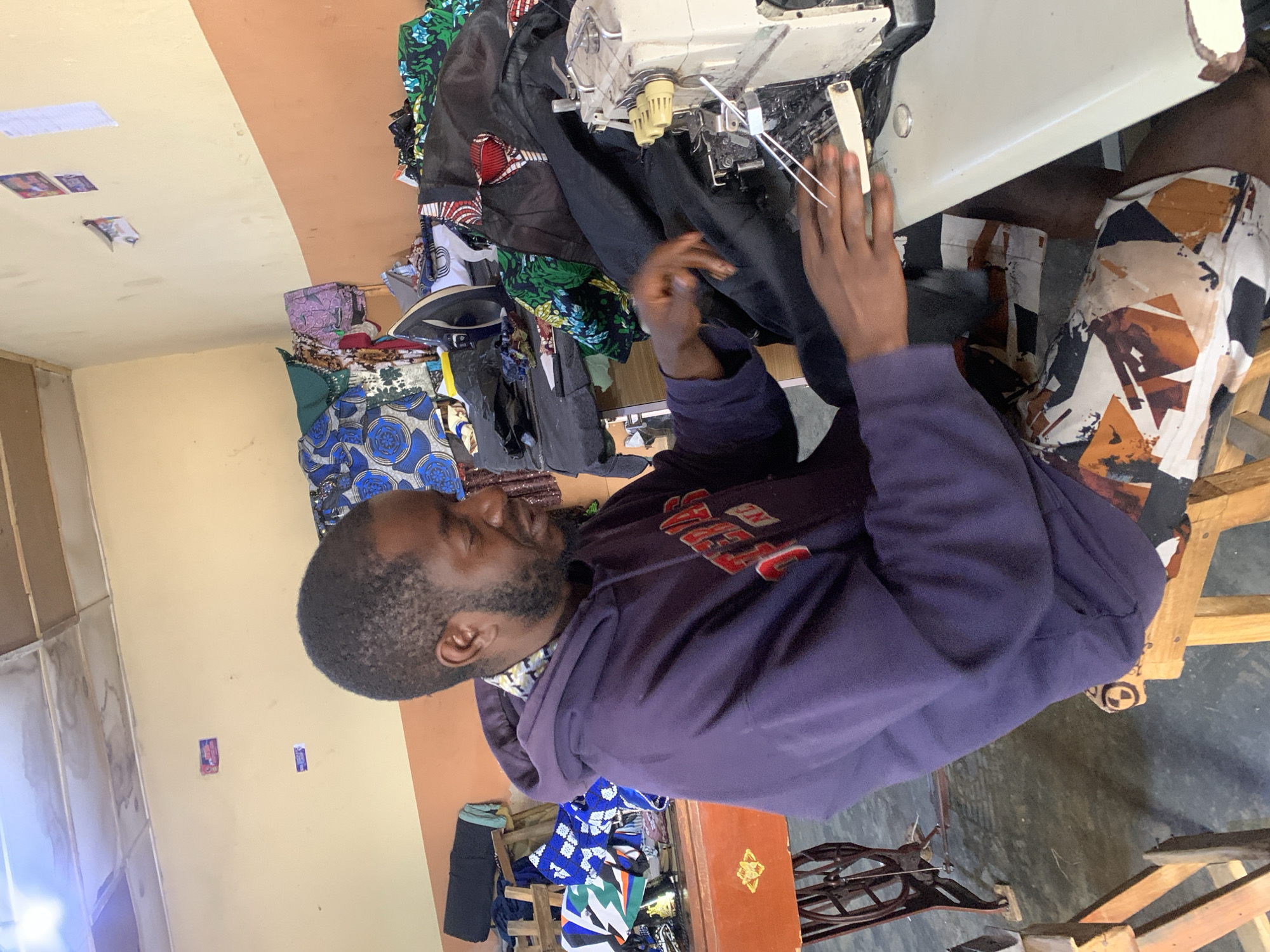 A man is seated at a sewing machine in Jos, Nigeria. He is wearing a purple hoodie and patterned shorts. The sewing machine is on a table, with various fabrics piled around it. The room has yellow and brown walls. Additional fabrics and a wooden stool are visible in the background.