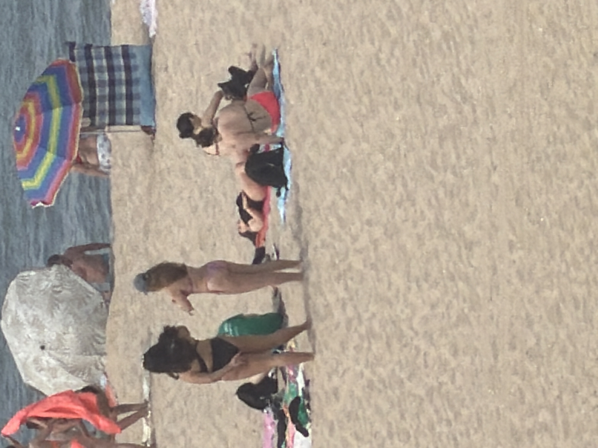 This image depicts a sunny day at a beach, likely in Espinho, Portugal, given the context. In the foreground, several individuals are visible on the sandy shore. Two women are standing, facing away from the camera, wearing bikinis. To their left, a woman in a black bikini stands, her back to the viewer. To her right, another woman in a light-colored bikini and a blue cap is also standing. In the mid-ground, several people are lying on towels or directly on the sand. One person is lying on their stomach on a red towel, wearing a dark bikini. Another person, wearing a black top and red bottoms, is lying on their back on a blue towel. Further back, two colorful beach umbrellas are set up, with a striped windbreak behind one of them. In the far background, individuals are visible near the water, and the ocean stretches to the horizon. Several beach towels and bags are scattered on the sand. The overall scene is one of leisure and recreation on a warm, clear day at the beach.