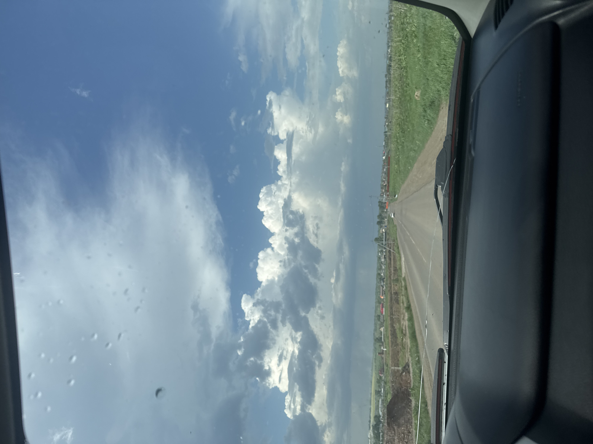 The image shows a view from inside a vehicle, likely a car or truck, looking out over a road and the surrounding environment. The sky dominates the view with a mix of blue sky and puffy white clouds. The road is straight and appears to be a highway with green fields on either side. A town with buildings and infrastructure is visible beyond the green fields. The perspective suggests the vehicle is in motion. The interior of the vehicle is visible on the right side of the frame, with the dashboard and part of the side window. The scene is outdoors, during daylight hours, with clear weather conditions but with some cloud cover. There's no evidence to suggest the city of Kitsjanovo.