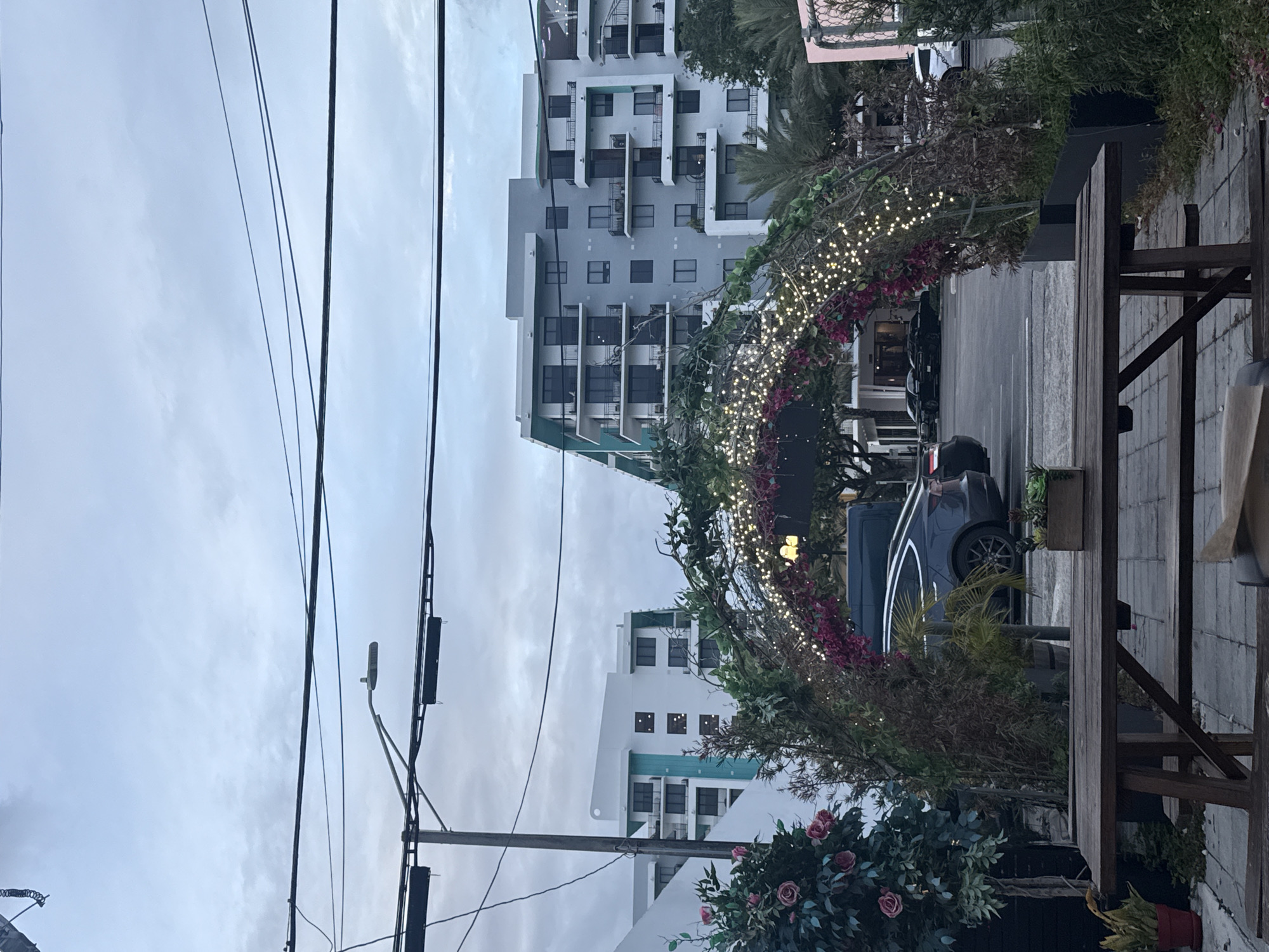 The image captures a static urban street scene under an overcast sky. In the foreground, several dark power lines diagonally traverse the upper half of the frame, connected to a utility pole on the left that also holds a street light. To the right, a wooden outdoor table and bench are partially visible, suggesting an outdoor seating area.
The midground is dominated by a decorative archway, elaborately adorned with green foliage, clusters of pinkish-red flowers, and numerous small, glowing string lights. This archway appears to span over a sidewalk or pedestrian path adjacent to a street where several cars are parked. Behind the archway and vehicles, multiple modern, multi-story buildings line the street. These buildings feature light grey facades, numerous windows, and balconies, with one prominent structure exhibiting a stepped architectural design and subtle teal accents. Various trees and bushes are interspersed throughout the scene, adding greenery.
The sky is a uniform light grey, indicating an overcast day. The illuminated string lights on the archway could suggest either dusk or evening, or simply a continuous decorative display. No human activity or specific event is taking place, and no discernible text is visible within the frame. The scene portrays a calm streetscape in a contemporary urban setting.