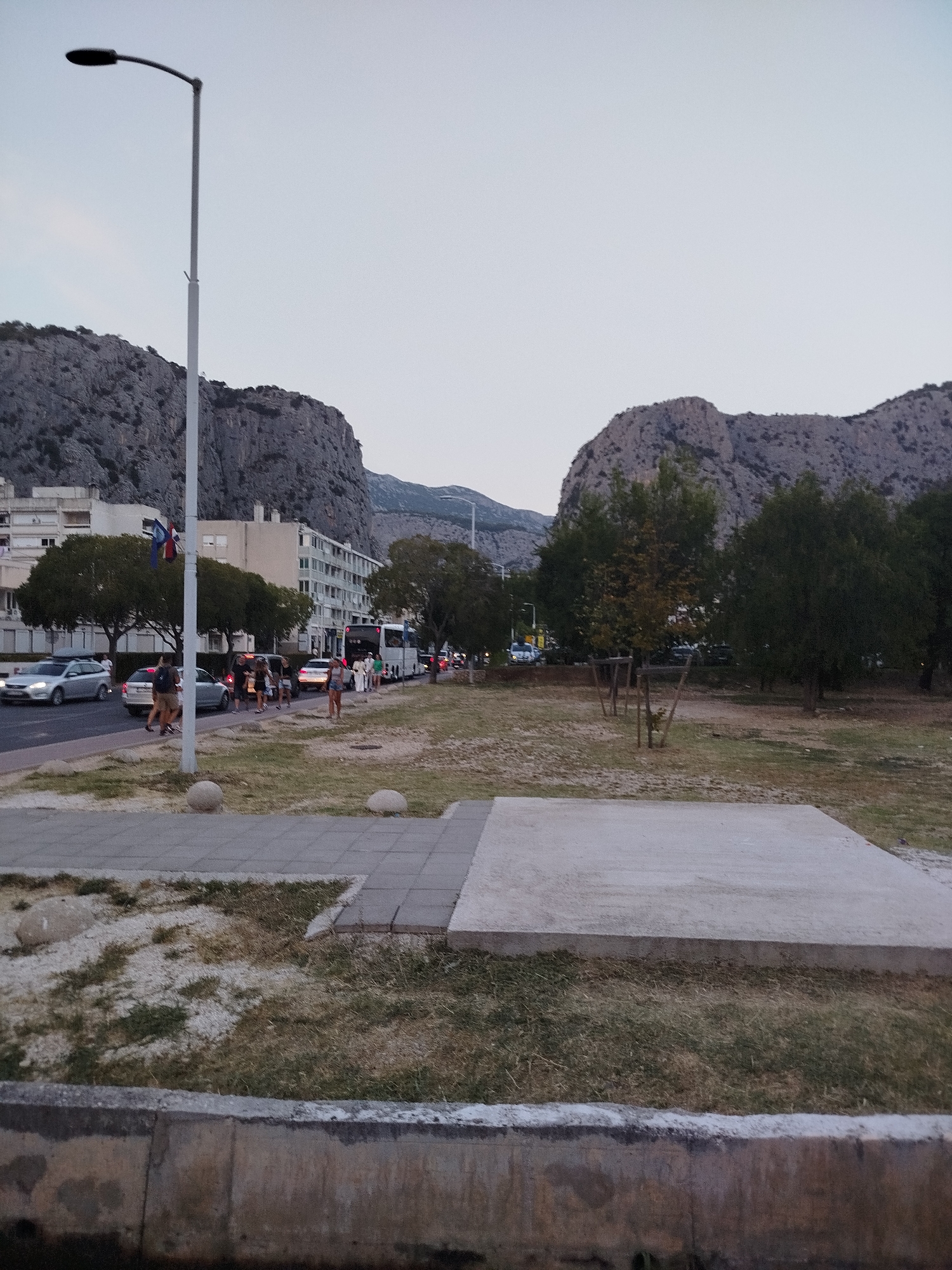 A street scene in Omiš, Croatia, features a road with several cars and a bus, suggesting moderate traffic. On the left side of the road, a sidewalk runs parallel to the street, with pedestrians walking along it. Behind the sidewalk is a large, rocky mountain range, a prominent natural feature of the area. Trees are sparsely planted along the road and in a grassy area to the right. A lamppost stands tall on the sidewalk, and two round concrete bollards are visible near the grassy area. The sky is overcast, indicating a cloudy day. The overall impression is an urban landscape integrated with natural surroundings.