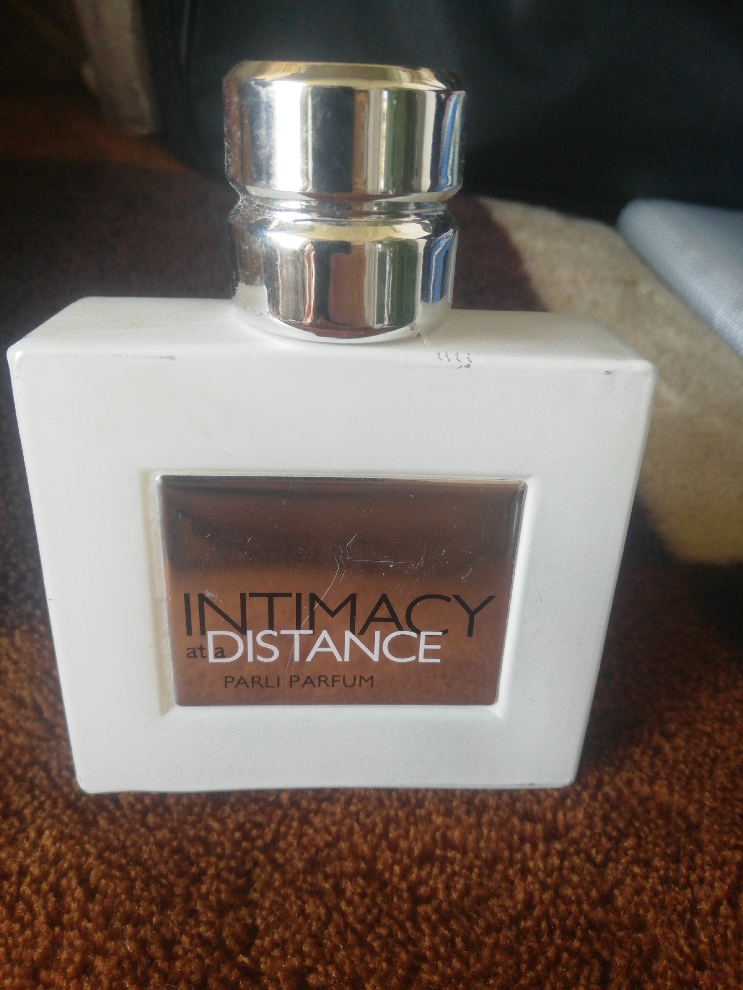 An overhead shot shows a white rectangular perfume bottle with a silver cap. The bottle has a dark brown label with white text. The text on the label reads "INTIMACY AT DISTANCE PARLI PARFUM". The bottle is placed on a brown fuzzy surface, possibly a rug. The background is dark and out of focus. There is no indication of location in the city of Unknown, Unknown.