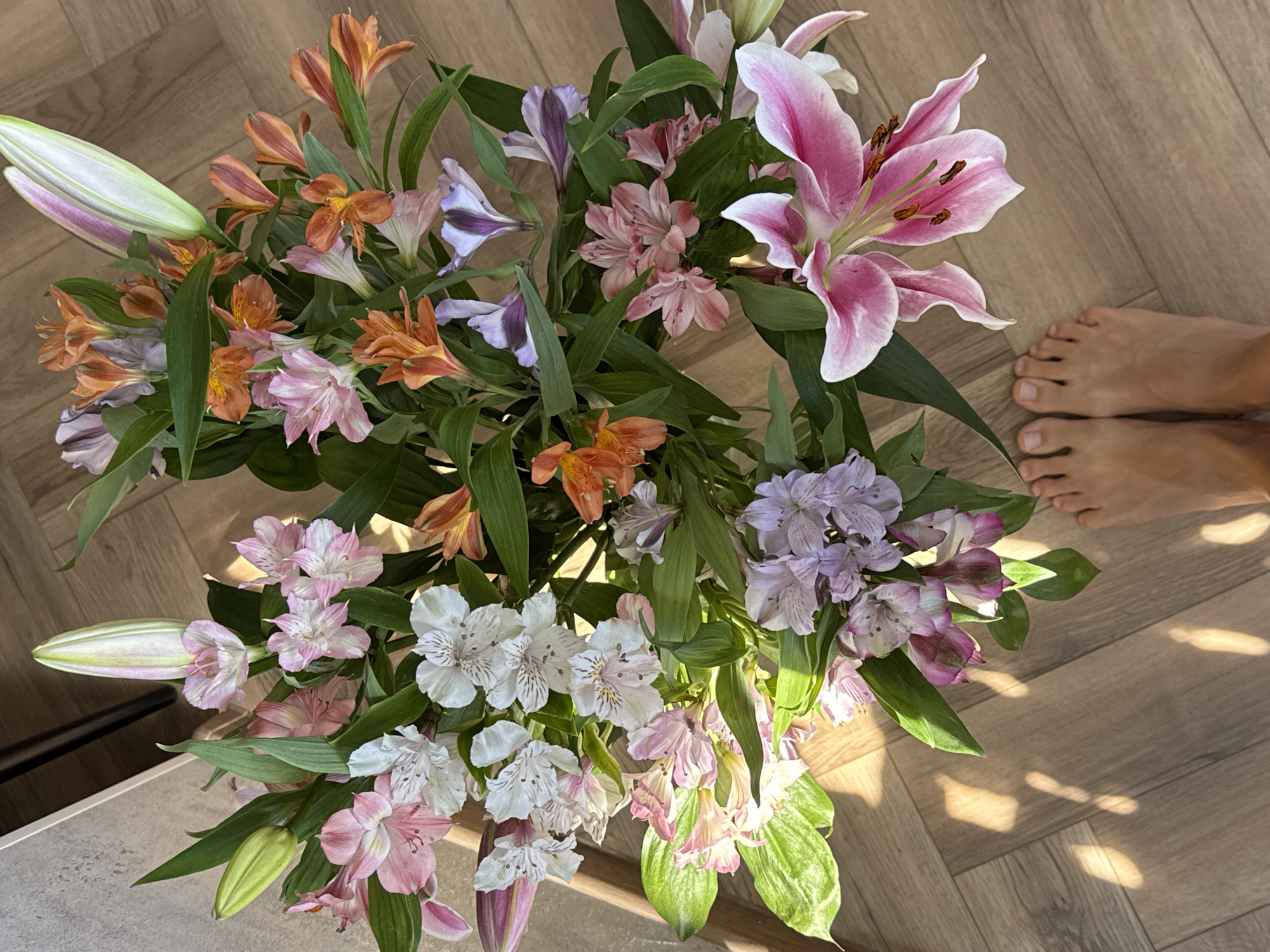 A high-angle shot depicts a bouquet of flowers placed on a light-brown wood-grain floor. The bouquet is composed of a variety of flowers including orange Alstroemeria, pink lilies, white Alstroemeria and purple Alstroemeria. The flowers are arranged with green foliage. Bare feet are partially visible in the top right corner of the image. Sunlight casts shadows across the floor. The location is Unknown, Unknown.