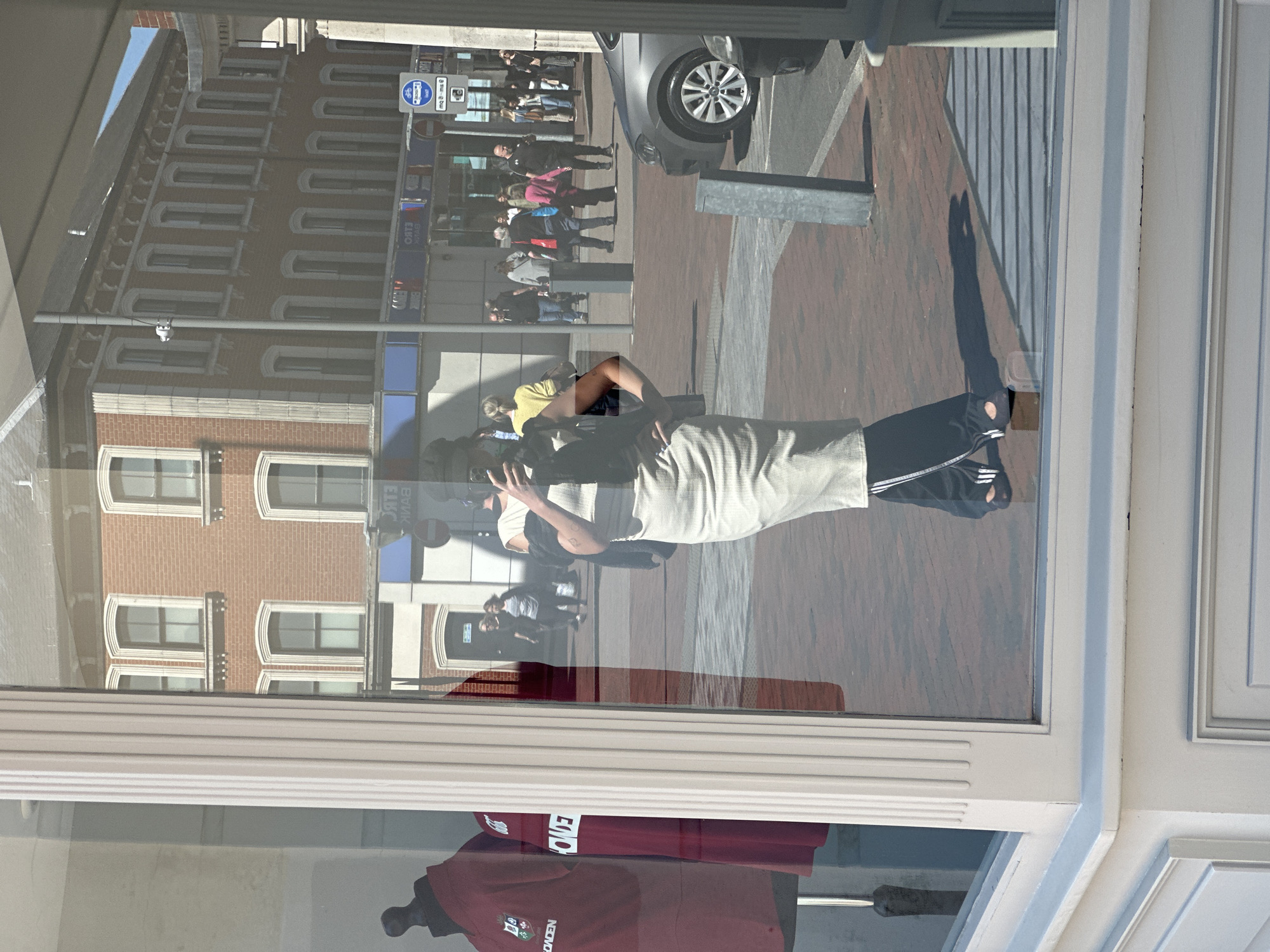 The image shows a window reflection in Tunbridge Wells, United Kingdom. The reflection captures a street scene with people walking, a silver car, brick buildings, and street signs. A woman holding a phone is reflected in the window. Below the reflection, a maroon shirt with the word "ROYDEN" is visible.
