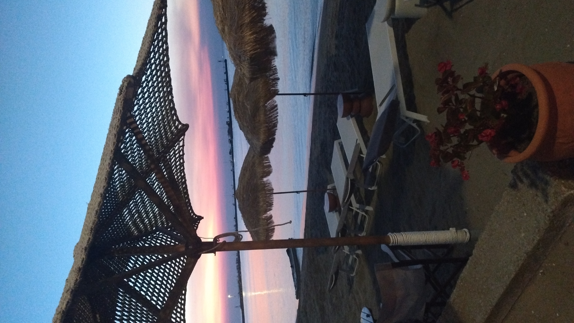 The image shows a beach at dusk in Neoi Epivates, Greece. The foreground features a large straw umbrella casting a shadow over several empty lounge chairs and more straw umbrellas lining the sandy beach. The sea is calm with a soft pink and purple sunset reflecting on its surface. In the distance, a pier or breakwater is visible, along with some lights. The overall atmosphere is peaceful and serene, indicating the end of a day at the beach.