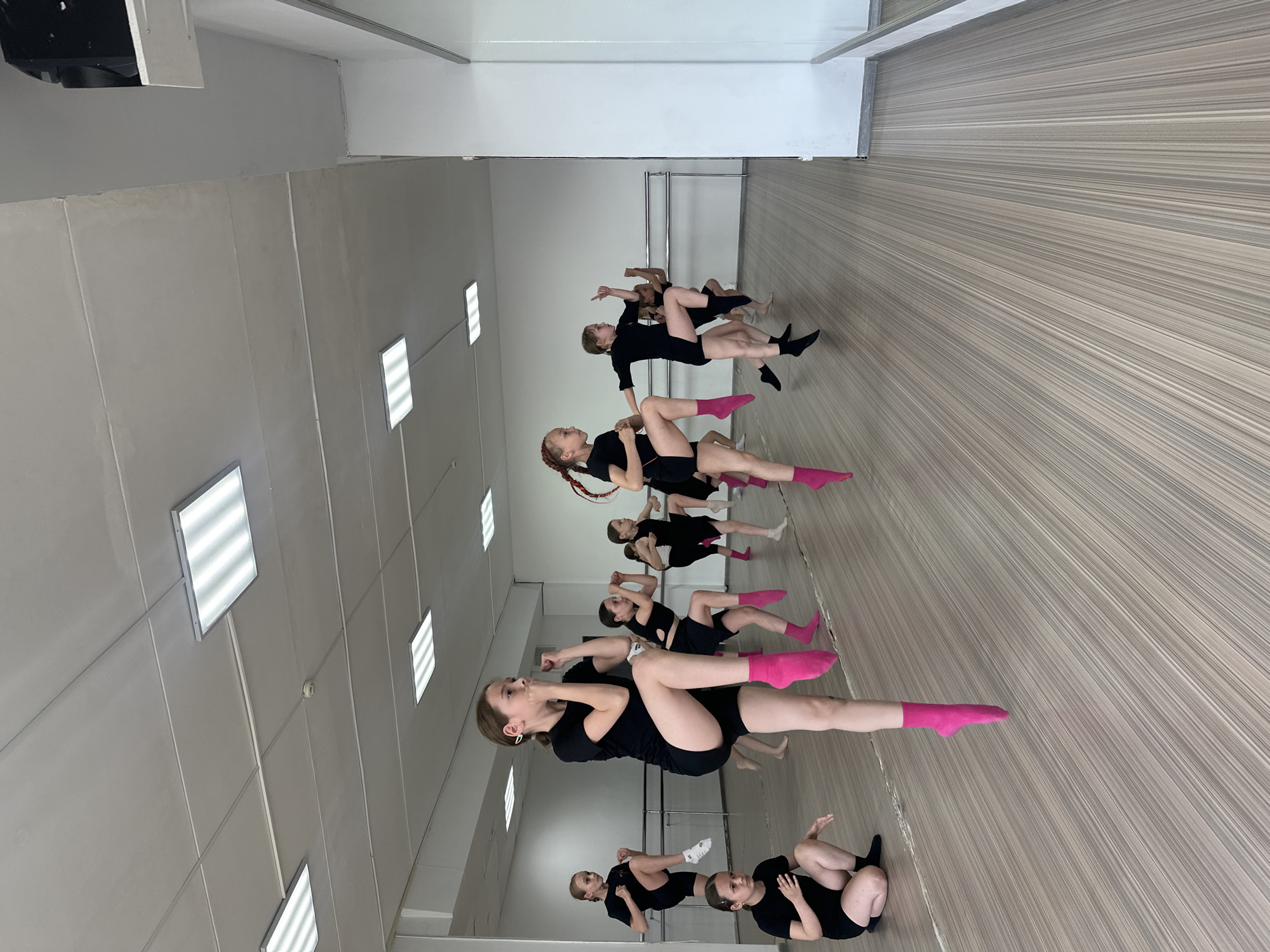 A dance class in what appears to be Votkinsk, Russia, as indicated by geotagging or related information. The dance studio has a mirrored wall and a light-colored, lined floor. Approximately 10-12 individuals, mostly young women, are participating. They are wearing black dance attire; some have on pink socks and others have on black socks. Some are in the middle of a routine or stretches on the floor. The walls are neutral-colored, with fluorescent lights on the ceiling. A ballet barre can be seen near the mirrored wall. The overall scene suggests a typical dance studio setting with students engaged in a class.