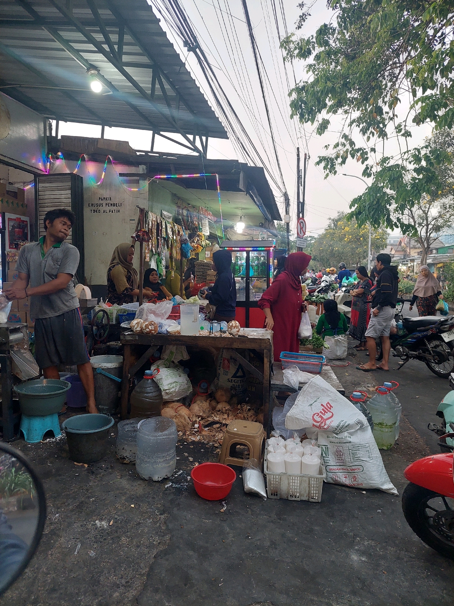 The image depicts a street-level market scene in Surabaya, Indonesia, with a focus on a vendor and customers. A man in a gray polo shirt and dark shorts stands to the left, appearing to be working with goods. In the background, several individuals are engaged in transactions or browsing. A woman in a maroon hijab stands near a stall, holding a plastic bag, and appears to be interacting with someone. Another woman in a darker hijab is visible behind her. The market is set up along a paved area, with various stalls and temporary structures. Goods such as coconuts, bags of sugar, and bottled water are visible. The environment suggests an outdoor market with temporary roofing, electrical wires overhead, and a tree branch extending into the frame. The overall atmosphere is that of a busy marketplace.
