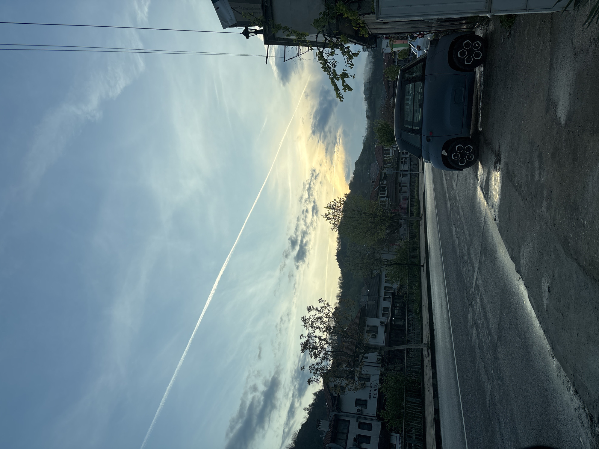 This media file captures a tranquil street scene in Zlatograd, Bulgaria, during either sunrise or sunset, indicated by the low angle of the sun casting a golden glow on the horizon and long shadows. The image is oriented vertically, showing a wide expanse of sky above a street lined with buildings and vegetation.
The sky, a clear light blue, is adorned with scattered white and grey clouds. A prominent, straight white contrail from an airplane cuts across the sky from left to right, subtly ascending. Two thin power lines are also visible in the upper sky.
In the foreground, a paved road stretches into the distance. It appears wet, reflecting the soft light from the horizon. A small, blue-grey electric microcar, possibly a Citroën Ami, is parked on the right side of the road, facing away from the viewer. Its distinctive wheels feature a multi-spoke design. The car and the immediate roadside are partially shrouded in shadow.
Along the street, several multi-story buildings with light-colored facades and red-tiled roofs are visible, interspersed with trees and bushes, some with vibrant green foliage, others still appearing somewhat bare. Air conditioning units are attached to some building exteriors. On one of the buildings, the Cyrillic text "КЪЩА ЗА ГОСТИ" (KASHTA ZA GOSTI - Guesthouse) and "ТОНИ" (TONI - Tony) is visible, suggesting a guesthouse named Tony. In the background, rolling hills or mountains are silhouetted against the bright sky, completing the serene landscape typical of the region. The overall impression is one of peacefulness in a quaint Bulgarian town.